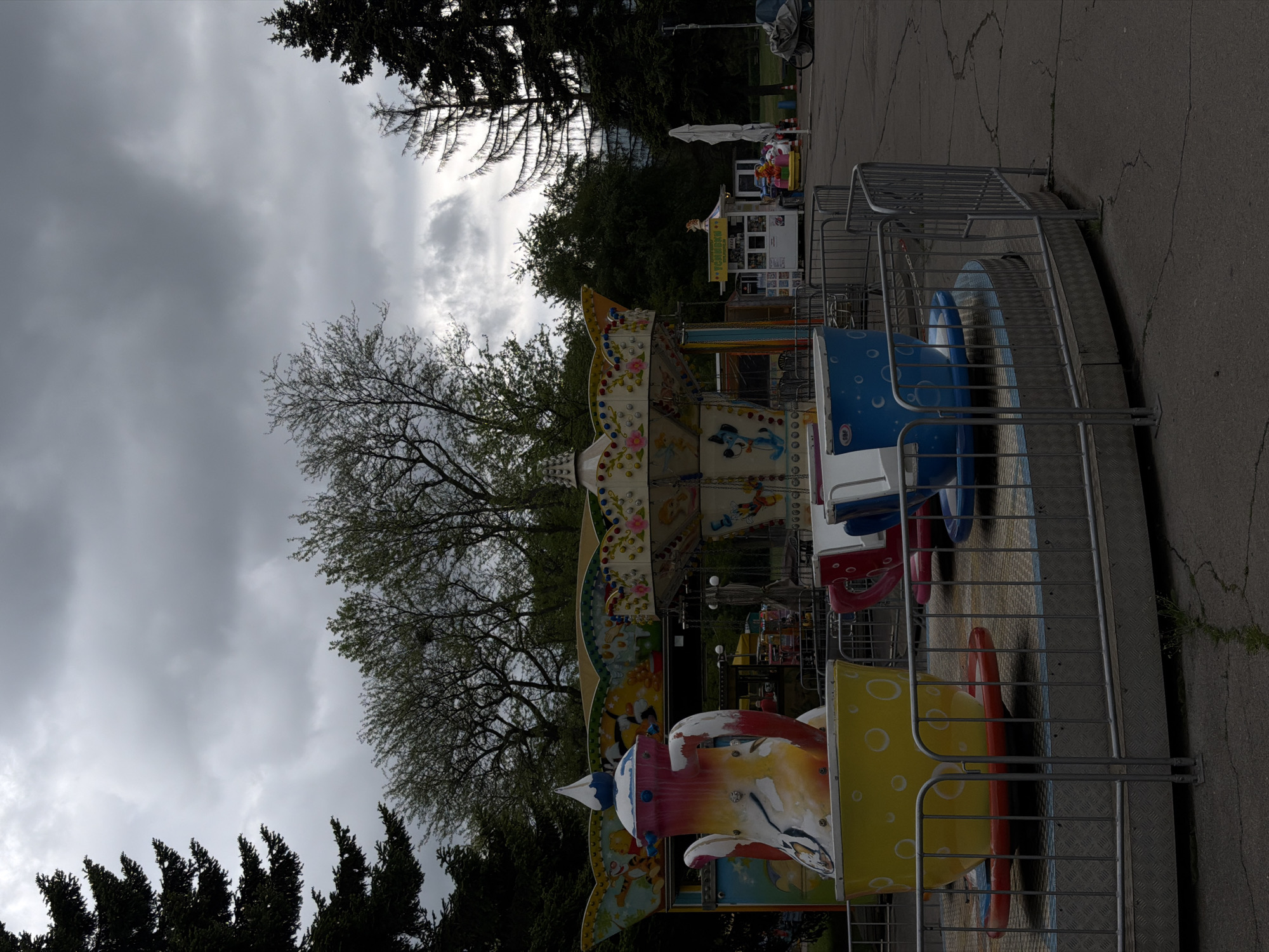 The image shows a children's amusement park ride located in Sofia, Bulgaria. The ride is a small carousel with colorful seats shaped like animals and objects. A gray metal fence surrounds the ride. The ground is paved and cracked. In the background, there are trees, cloudy skies, and a small building that is possibly a ticket booth.