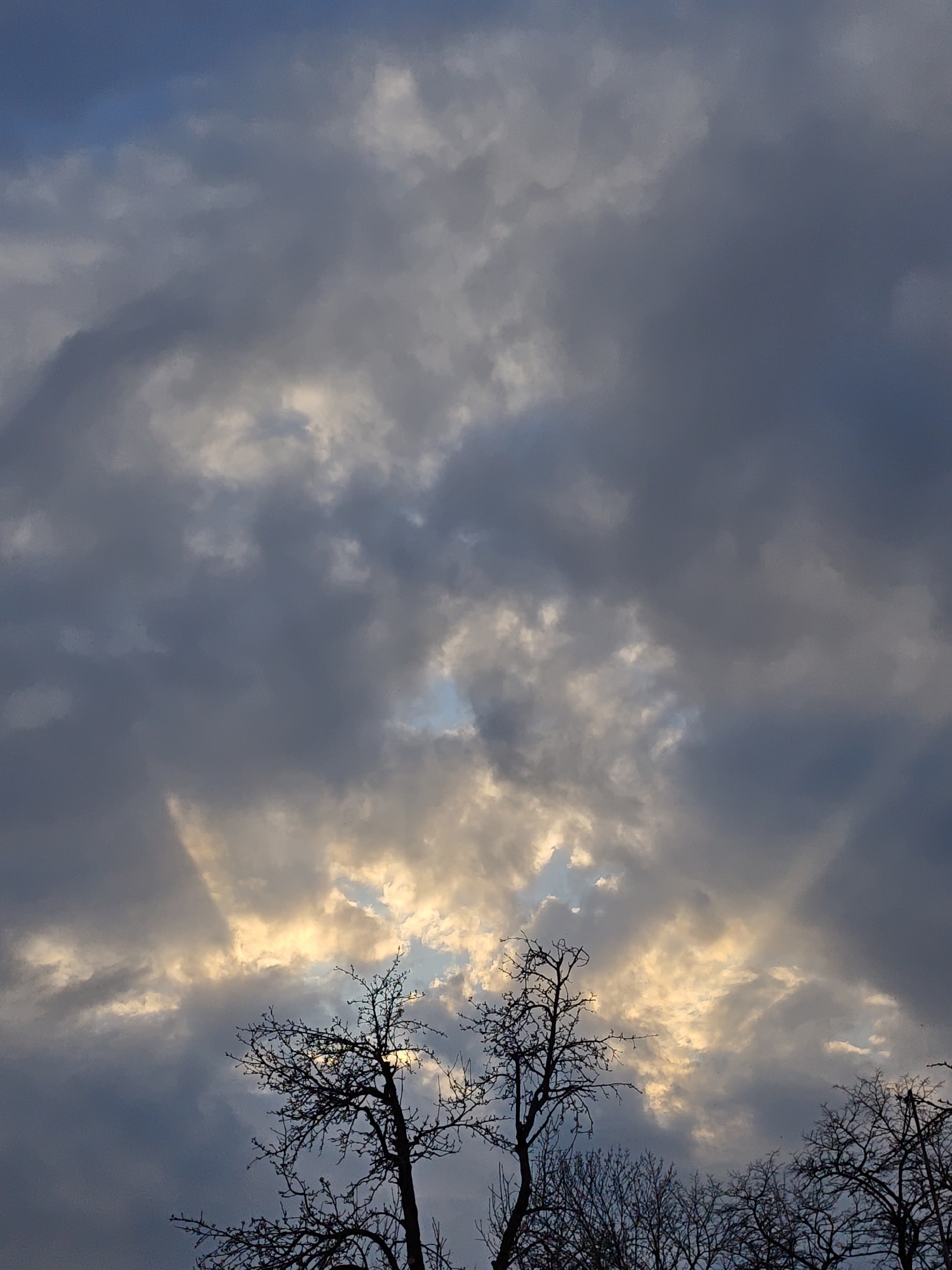 The image shows a view of the sky with dark, heavy clouds covering most of the frame. There are breaks in the clouds through which sunlight shines, creating a bright glow. In the lower portion of the image, silhouetted bare trees are visible against the backdrop of the sky. The scene appears to be a natural setting, possibly a park or rural area. The location is Parkhomivka, Ukraine.