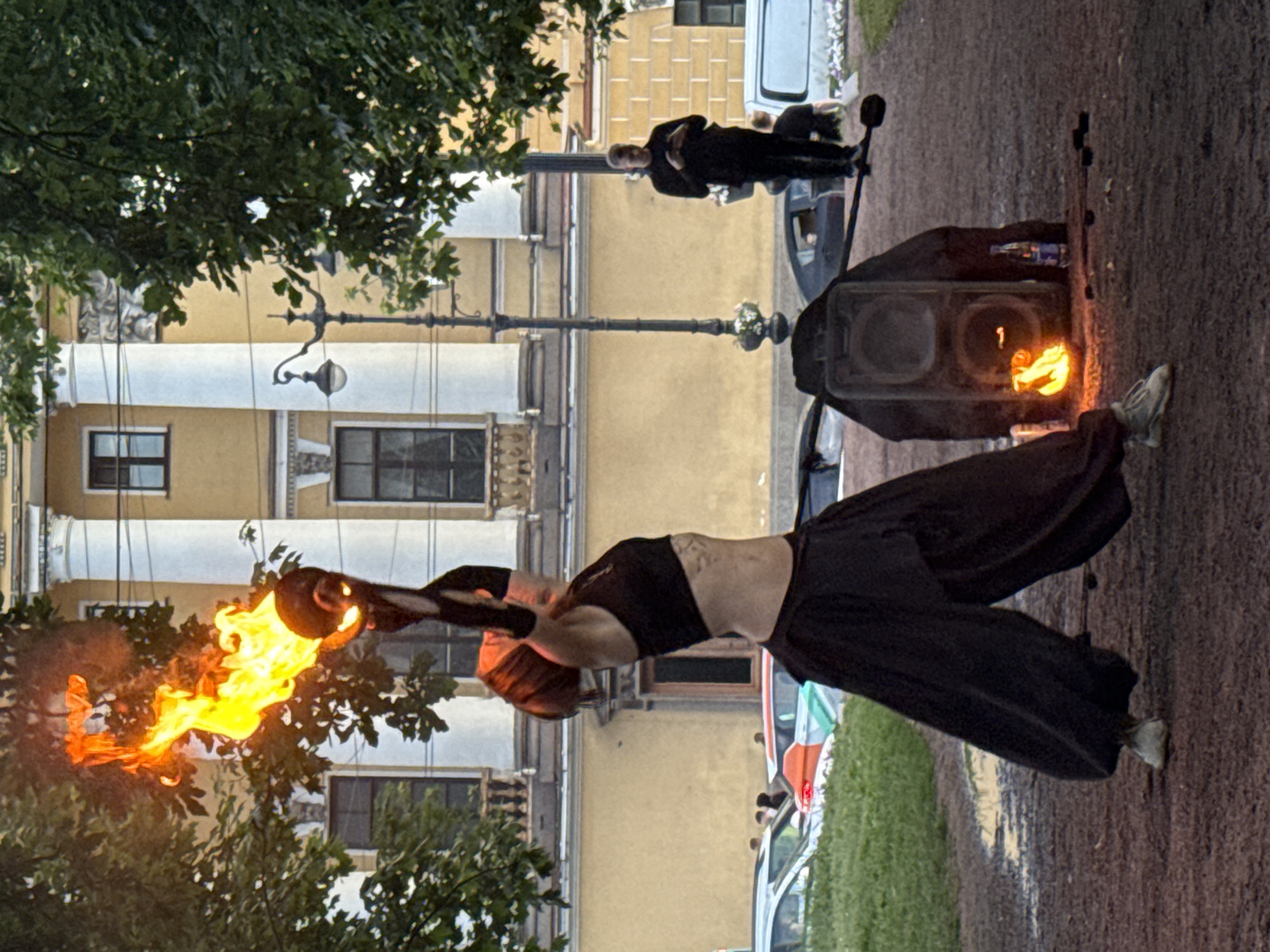 A person is performing fire-spinning in front of a building with columns in Saint Petersburg, Russia. The performer, dressed in a black crop top and wide black pants, is holding a flaming prop above their head. The background features a yellow building with classical architecture and large trees. A street lamp and a speaker are visible on the ground. Several cars and a few people are in the background, suggesting a public outdoor space. The ground appears to be a mix of dirt and wet patches, possibly indicating recent rain.