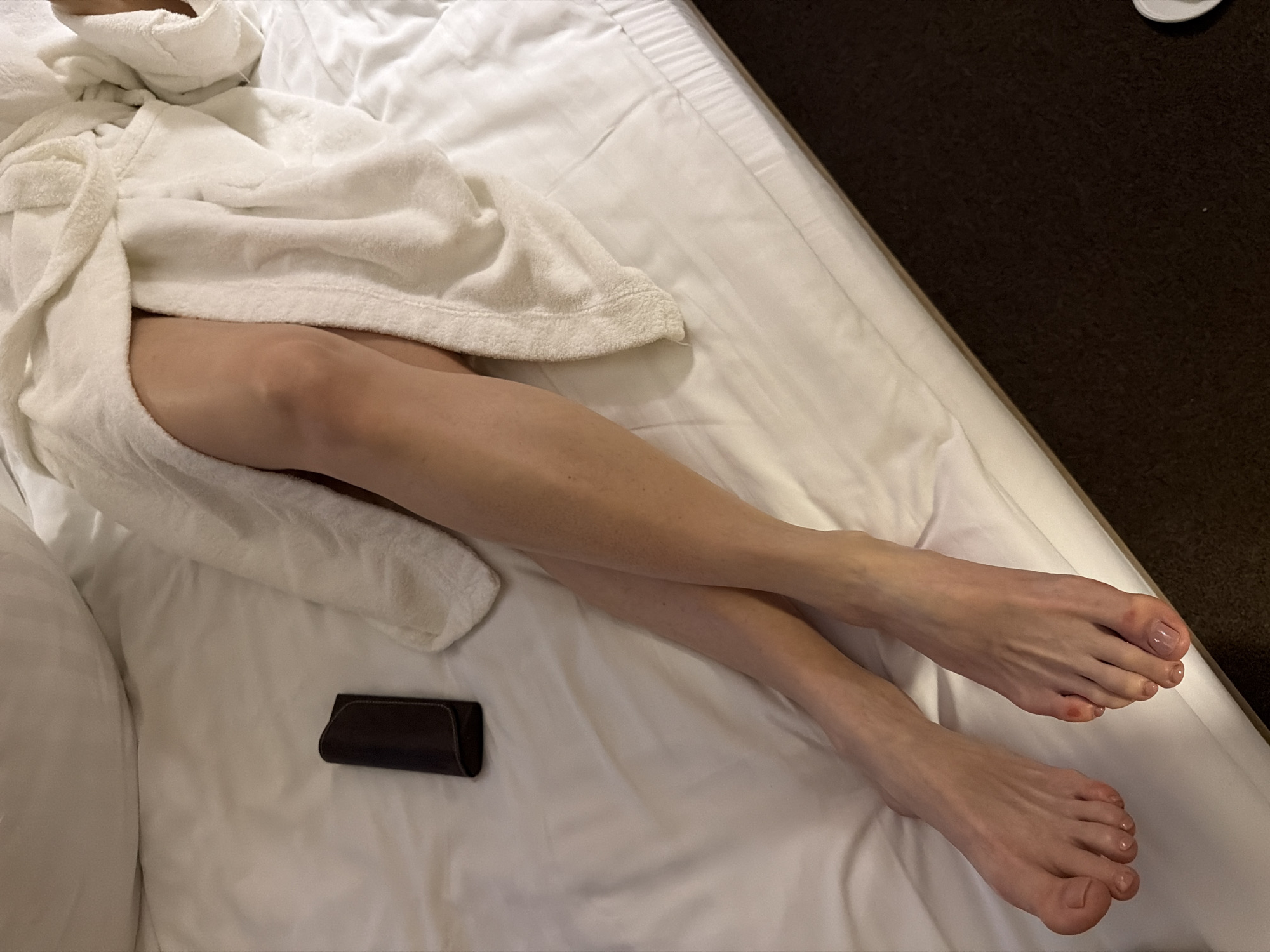 The image presents an overhead view of a person reclining on a white-sheeted bed. The individual, whose gender is not explicitly identifiable but appears to be an adult, wears a white terry cloth bathrobe, which is partially open. Both legs are visible from mid-thigh down, extending towards the bottom of the frame, with the feet slightly crossed at the ankles. The toes exhibit a clear or very pale pink nail polish. To the left of the person's legs, a dark brown, rectangular object, possibly a glasses case or small pouch, rests on the white bedding. The bed features clean, white sheets and a large white pillow partially visible at the top. The bottom and right edge of the frame reveal a dark brown carpeted floor. The setting appears to be an indoor environment, consistent with a bedroom or hotel room. The image itself provides no identifying information regarding the specific geographic location, such as the city of Svetlogorsk, Russia.