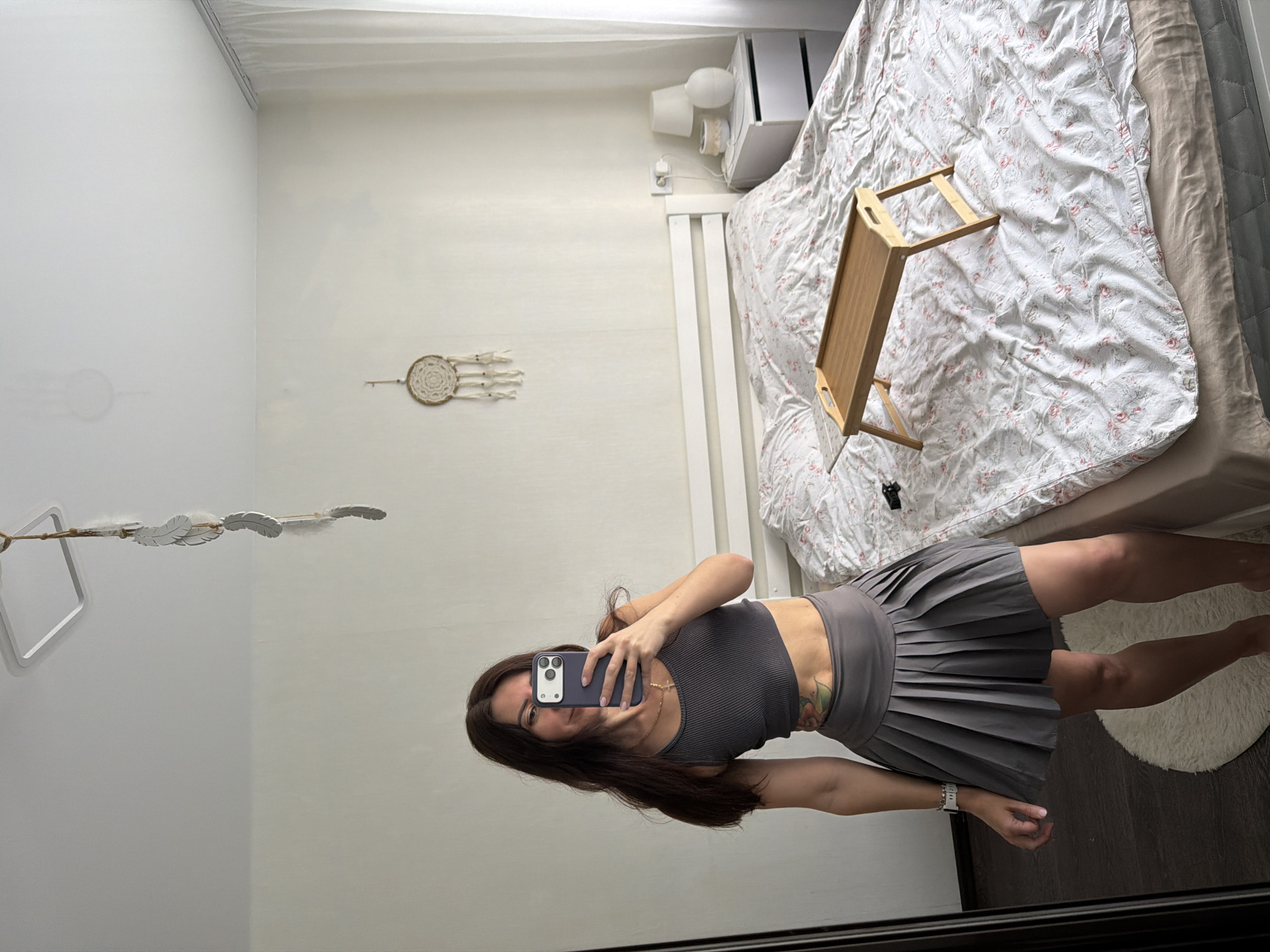 A woman is taking a mirror selfie in a bedroom. She is standing in front of a large mirror, holding a smartphone in her right hand. She is wearing a gray crop top and a gray pleated skirt. Her hair is dark brown and falls past her shoulders. She has a tattoo on her stomach. The bedroom is minimally decorated. A bed with a floral patterned duvet is visible behind her. A small wooden tray table is placed on the bed. A nightstand with a lamp and other objects is next to the bed. A white curtain is visible on the right side of the room. A dreamcatcher hangs on the wall above the bed. The overall impression is a casual, personal space.