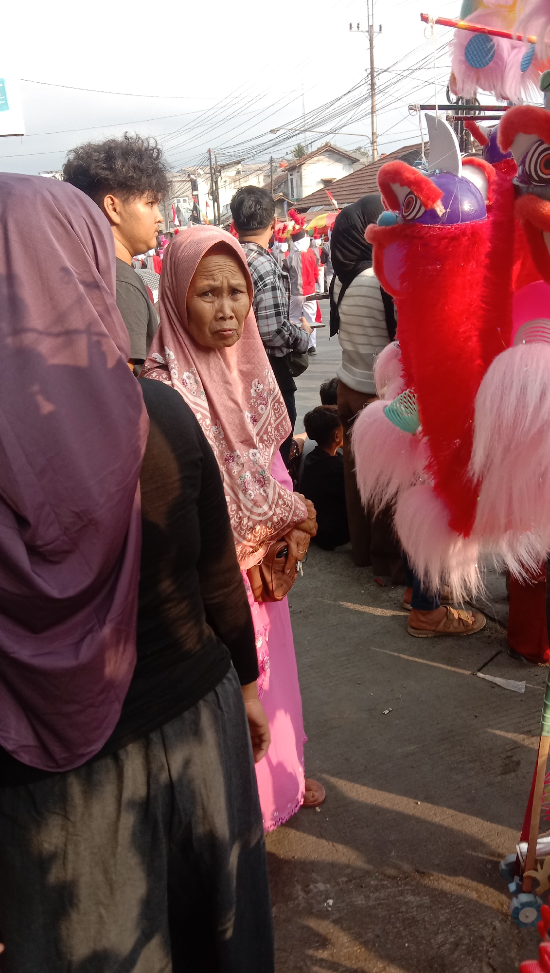 An outdoor medium shot captures a group of people in what appears to be a street or public gathering. In the foreground, an older woman wearing a pink hijab and a matching patterned dress is looking directly at the camera with a somewhat stern expression. She is holding a small brown purse. To her left, a person wearing a purple hijab and a black top with a grey skirt is partially visible. Further back, a young man with curly hair is looking to his left, and other individuals in the background are observing a parade or event, some wearing red and white uniforms. Colorful, oversized toy-like figures, possibly part of a procession or market stall, are prominent on the right side of the frame, featuring bright red and pink fur. The environment suggests a sunny day with some power lines and buildings visible in the distance.
