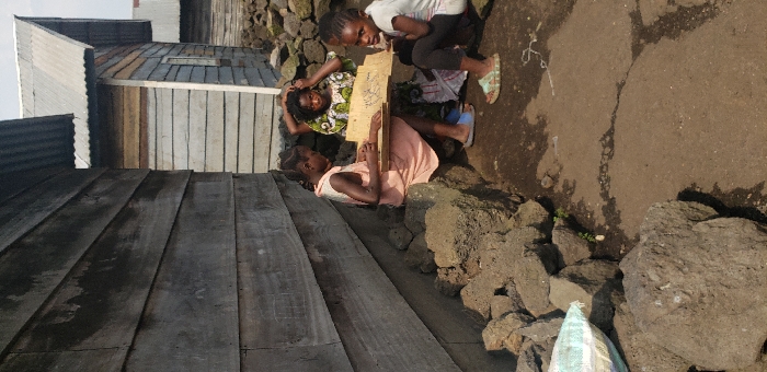 The image depicts an outdoor scene in Buzove, Democratic Republic of the Congo, featuring three individuals near two wooden-plank structures with corrugated metal roofs under clear daylight. In the foreground, the ground is composed of compacted dirt with visible cracks and numerous dark, irregularly shaped rocks.
From left to right, a young female in a light-colored dress sits on a pile of these rocks, holding a rectangular wooden board with visible markings on its surface. She appears engaged with the board, looking down. Her feet are shod in blue slide sandals. Behind her, an older female or youth, dressed in a patterned garment featuring green, white, and yellow hues, sits in a slightly elevated position, with her hands near her head. To the right, another young female, wearing a white short-sleeved top and dark pants, sits directly on the dirt ground, looking towards the viewer. She wears patterned slide sandals.
On the far left, a portion of a white and blue patterned sack is visible on the ground. The building immediately to the left is constructed of weathered, horizontal wooden planks, with a corrugated metal roof. Another similar wooden building with a corrugated metal roof is visible behind and to the right of the individuals. The overall environment suggests a residential or community area.