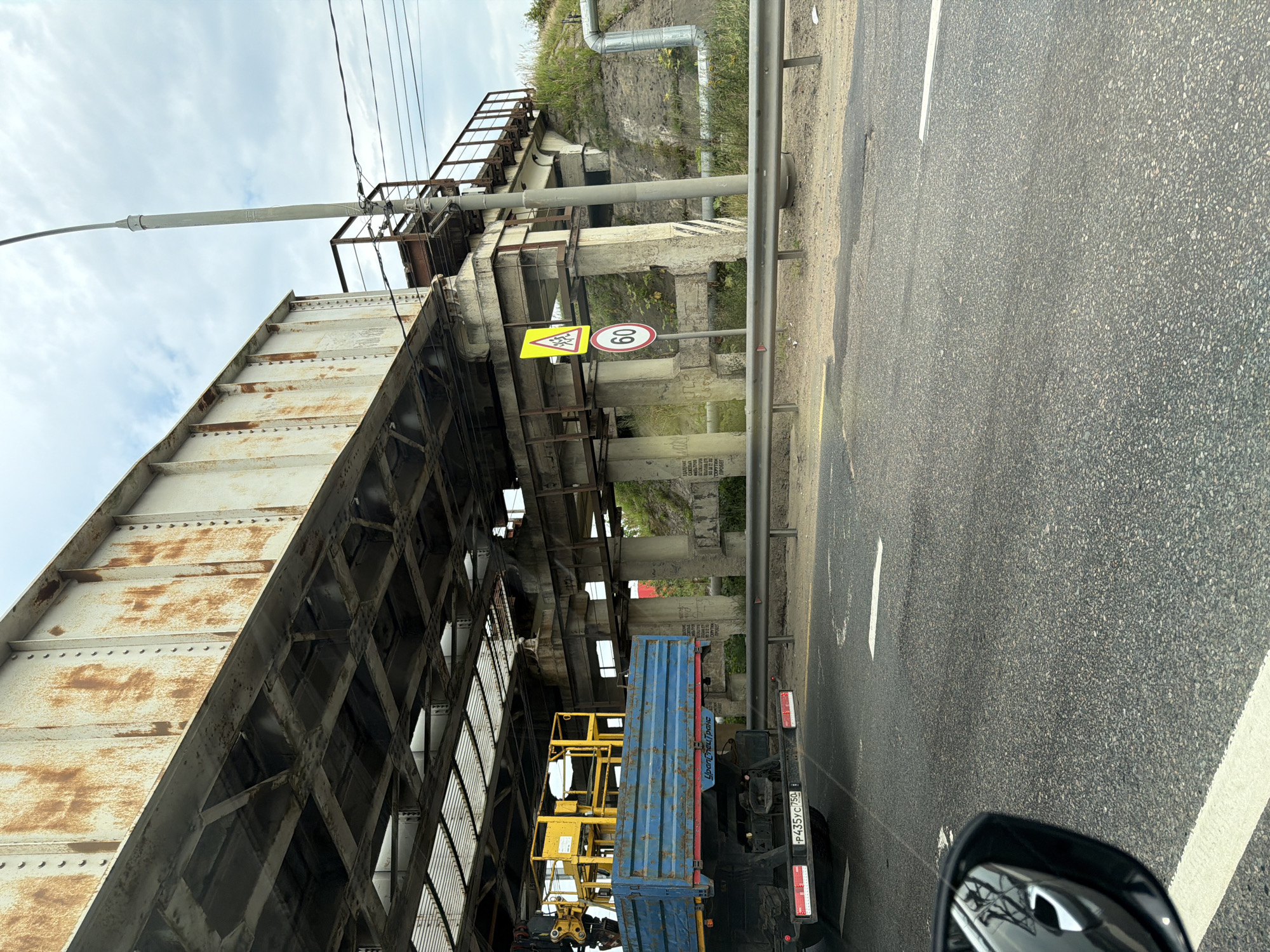 The image depicts a view from a vehicle driving on a paved road, passing under an old, rusty railway bridge. To the right of the road, there is a metal guardrail. A truck with a blue flatbed is parked partially under the bridge on the left side of the frame. The truck has a yellow aerial work platform on its bed and a license plate that reads "P435YC 750". A traffic sign with a yellow triangle depicting children crossing is visible, followed by a circular speed limit sign showing "60". A tall lamppost stands to the right of the bridge. Concrete pillars support the bridge structure, and there is overgrown grass and foliage on the embankment behind the guardrail. Pipes are visible running along the embankment. The sky is overcast with clouds. The scene appears to be located in Radumlya, Russia, based on the provided context, likely on a road near or within the vicinity of the town's infrastructure. There are no people clearly visible in the image.