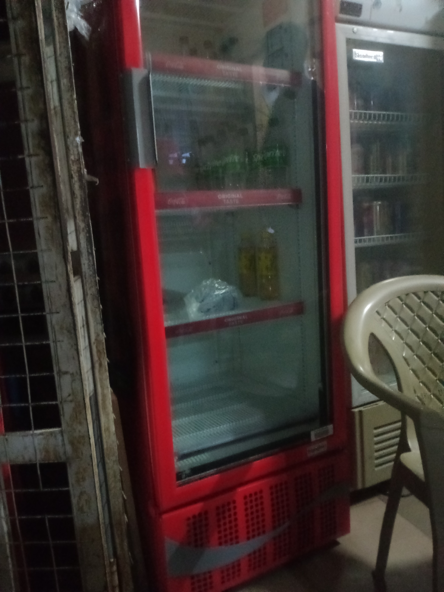 In Jalingo, Nigeria, a red Coca-Cola branded beverage cooler is prominently displayed, its glass door partially open, revealing shelves stocked with bottles of drinks and a bag of what appears to be ice. To the left of the cooler, a metal cage-like structure, possibly a gate or part of a shop's exterior, stands, showing signs of age and wear. Adjacent to the Coca-Cola cooler, another, older-looking white cooler with multiple shelves is visible, with various cylindrical containers on its shelves. In the foreground, the back of a light-colored plastic chair with a patterned seat is captured, suggesting a seating area nearby. The overall environment appears to be an indoor space, possibly a shop or an outdoor stall, with dim lighting contributing to a somewhat cluttered and aged aesthetic.