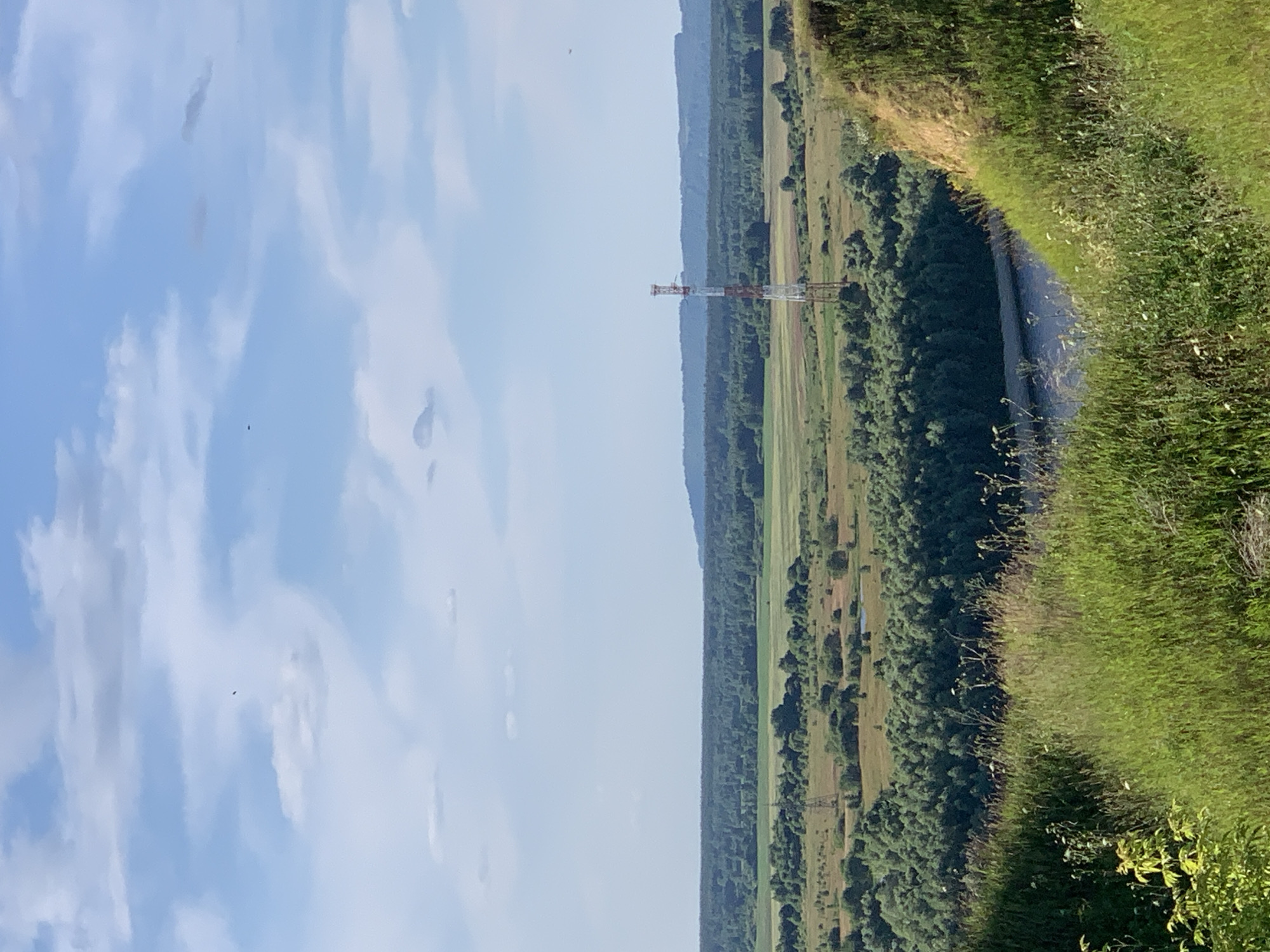 This is a vertical, landscape-oriented photograph taken outdoors on a clear, bright day. The scene depicts a wide, sweeping view of a rural landscape, likely in Pavlovo, Russia. The foreground is dominated by a grassy embankment on the right side, sloping downwards towards a body of water that appears to be a river or a large stream. The water is dark and reflects the sky.
Beyond the water, the landscape opens up to a vast expanse of green fields and dense forestation. Patches of lighter green suggest agricultural fields interspersed with darker, more textured areas of trees. In the middle ground, a tall, red and white lattice tower, likely a telecommunications or power pylon, stands prominently against the horizon. The horizon itself is marked by a series of undulating, blue-hazed hills or mountains, fading into the distance.
The sky occupies the left portion of the frame and is a bright, pale blue with scattered white, wispy clouds. There are no people visible in the image. The overall impression is one of tranquility and natural beauty, with the man-made tower being the only significant structure breaking the natural lines of the landscape. The lighting suggests it is daytime, likely mid-morning or afternoon, with the sun high in the sky, casting minimal shadows. The weather appears to be pleasant and clear. There is no visible text in the image.