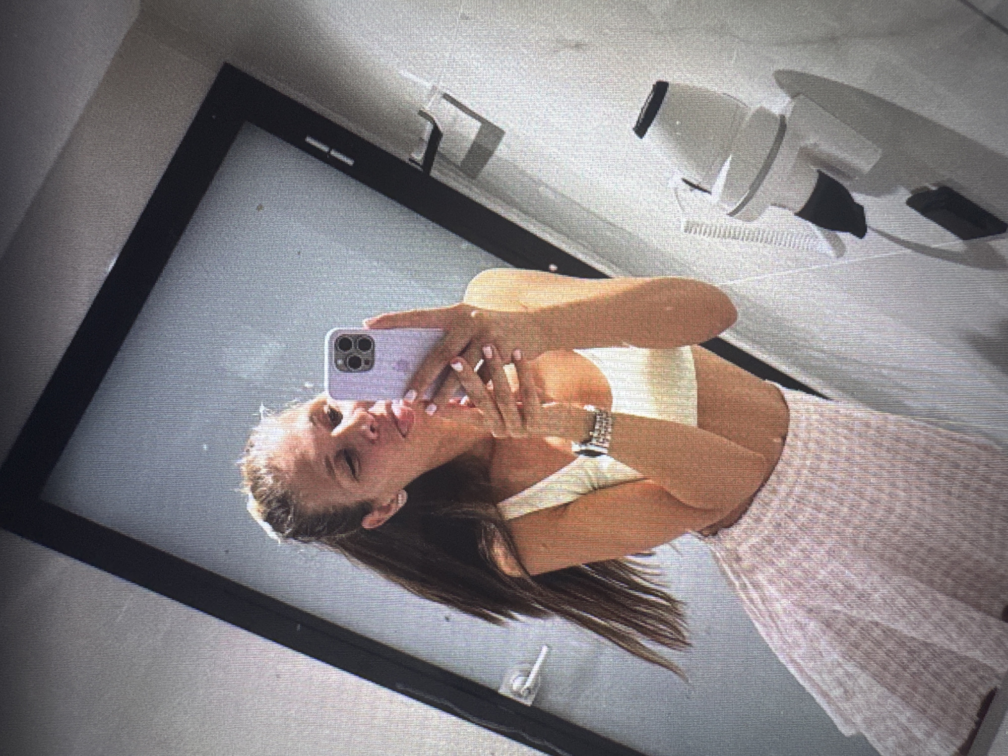 An eye-level selfie photograph captures a young adult female reflected in a mirror. The female, positioned centrally, has long, dark brown hair styled in a high half-up or ponytail. She wears a light-colored, possibly cream or white, sleeveless crop top that exposes her midriff, and a light pink pleated mini skirt. A silver-toned smartwatch with a link band is visible on her left wrist, and her fingernails are painted light pink. She holds a light purple-cased smartphone with three visible camera lenses, obscuring most of her face, but her eyes appear closed or squinted and her tongue is slightly extended in a playful gesture.
The background, seen as a reflection in the mirror, suggests an indoor setting consistent with a public restroom, changing room, or private vanity area. To the left of the female, a dark-framed door with a silver-toned lever handle is visible. To her right, a light-colored wall features a wall-mounted white hairdryer with a black nozzle and coiled cord, situated above a dark, possibly black, mounting bracket or fixture. Another dark, L-shaped hook or bracket is mounted higher on the same wall segment. The mirror itself has a dark, possibly black, frame, and the reflective surface shows minor imperfections. The lighting appears artificial. The image contains no discernible features or text to specifically identify the location within the city of Vratsa, Bulgaria.