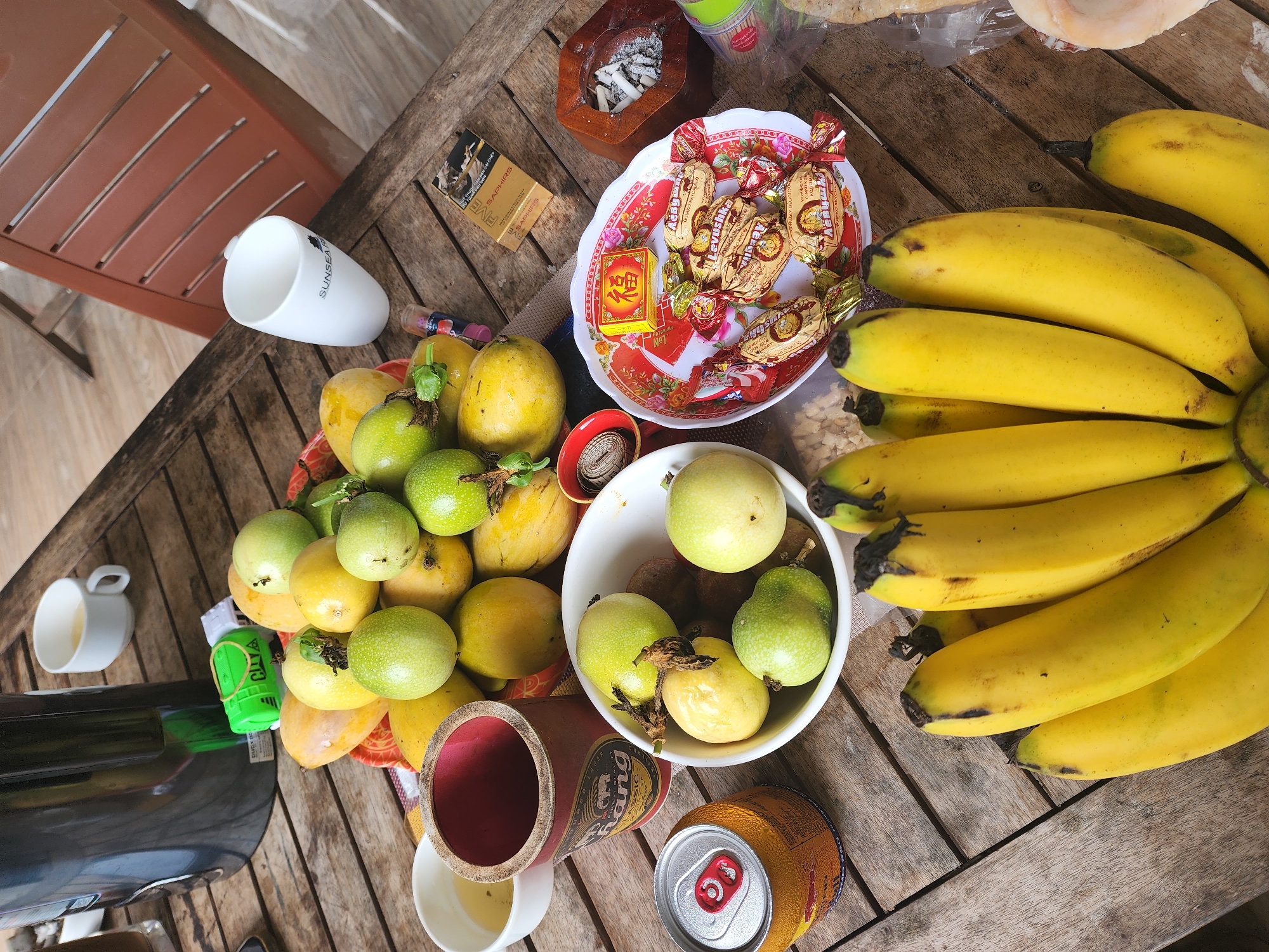 The image depicts a close-up, high-angle view of a wooden table laden with various fruits, drinks, and smoking accessories. The setting appears to be outdoors, possibly a patio or balcony, as suggested by the wooden slatted chair visible in the background and the natural lighting. The table itself is made of aged, weathered wood planks, creating a rustic ambiance.
Dominating the center of the frame is a large assortment of passion fruits and mangoes, piled in a basket and a white bowl. The passion fruits exhibit a gradient of green and yellow, indicating ripeness, while the mangoes are a vibrant yellow. Surrounding the fruits are several beverage containers: a tall, dark thermos, a red and yellow soda can, and two white ceramic mugs, one containing a light-colored liquid.
To the right of the fruit display, a small, ornate plate holds a collection of individually wrapped candies, some with gold foil wrappers featuring red accents. A yellow box with Chinese characters sits atop the candies, possibly a good luck charm or festive decoration. Adjacent to the plate is a wooden ashtray overflowing with cigarette butts and ash, indicating recent smoking activity. A pack of "Saphirs" cigarettes lies nearby.
In the foreground, a bunch of ripe yellow bananas is prominently displayed. Scattered amongst the items are other smaller objects, including what appears to be a small toy, and a red circular object that might be a bottle cap or lid. The overall impression is one of a relaxed, informal gathering or break, with an emphasis on fresh produce and leisure items. The scene is devoid of any identifiable individuals, focusing solely on the arrangement of objects on the table. The image does not contain specific indicators of location within Phan Thiet Ward, Vietnam, beyond the general cultural context suggested by the presence of what appear to be local snacks and potentially regional fruit varieties.