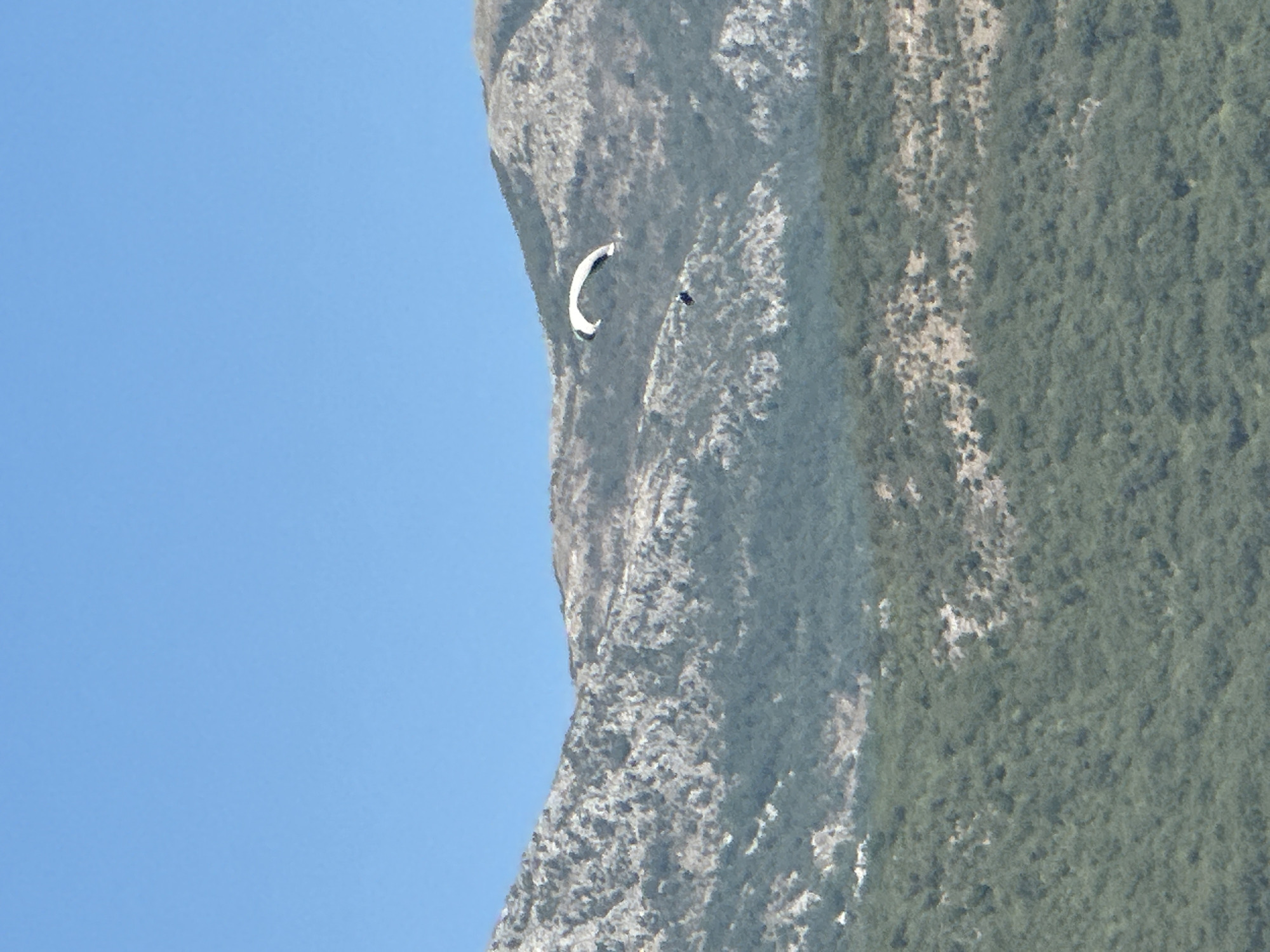 A paraglider is soaring through the sky above a mountain range. The paraglider, with a white canopy and dark trim, is visible in the upper right quadrant of the image. Below the paraglider, a second, smaller dark figure, likely the paraglider pilot, is suspended beneath the canopy. The mountains in the background are rugged and covered with sparse vegetation, transitioning from rocky, lighter gray slopes at the top to denser green foliage lower down. The sky above is a clear, unclouded blue. The scene appears to be a scenic outdoor location, possibly near Bečići, Montenegro, a region known for its mountainous terrain and coastal views suitable for paragliding.