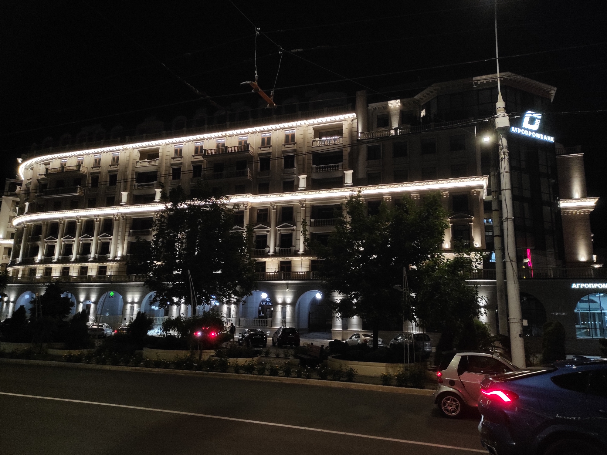 The image shows a nighttime street scene in an urban location. Prominently featured is a large, well-lit building with architectural details such as columns, balconies, and arches. The building's signage includes the name "АГРОПРОМБАНК" in Cyrillic. The street is lined with trees and shrubs, and several cars are visible, including a silver Smart car and a dark-colored vehicle in the foreground with illuminated taillights. Streetlights provide additional illumination. A utility pole with wires is also visible.