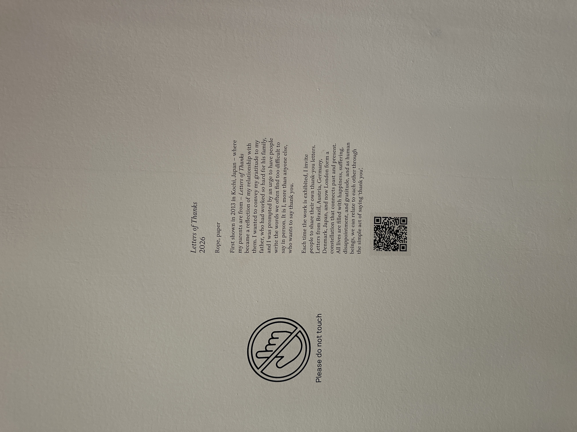 The image is of a descriptive card placed next to an art installation. The card reads "Letters of Thanks" with the medium listed as "Rope, paper" and the year as "2026". It provides context for the artwork and indicates that the work was first exhibited in Kochi, Japan, in 2013, where the artist's parents are from. The text discusses themes of gratitude and the difficulty of expressing thank yous. The card also states that the artist invites people to share their thank-you letters when the work is exhibited, and these letters form a constellation connecting various countries, including Brazil, Austria, Germany, Denmark, Japan, and now London. Below the text is a QR code and an icon of a hand with a line through it and the text, "Please do not touch." The mention of London within the card indicates that the artwork is currently being exhibited there.