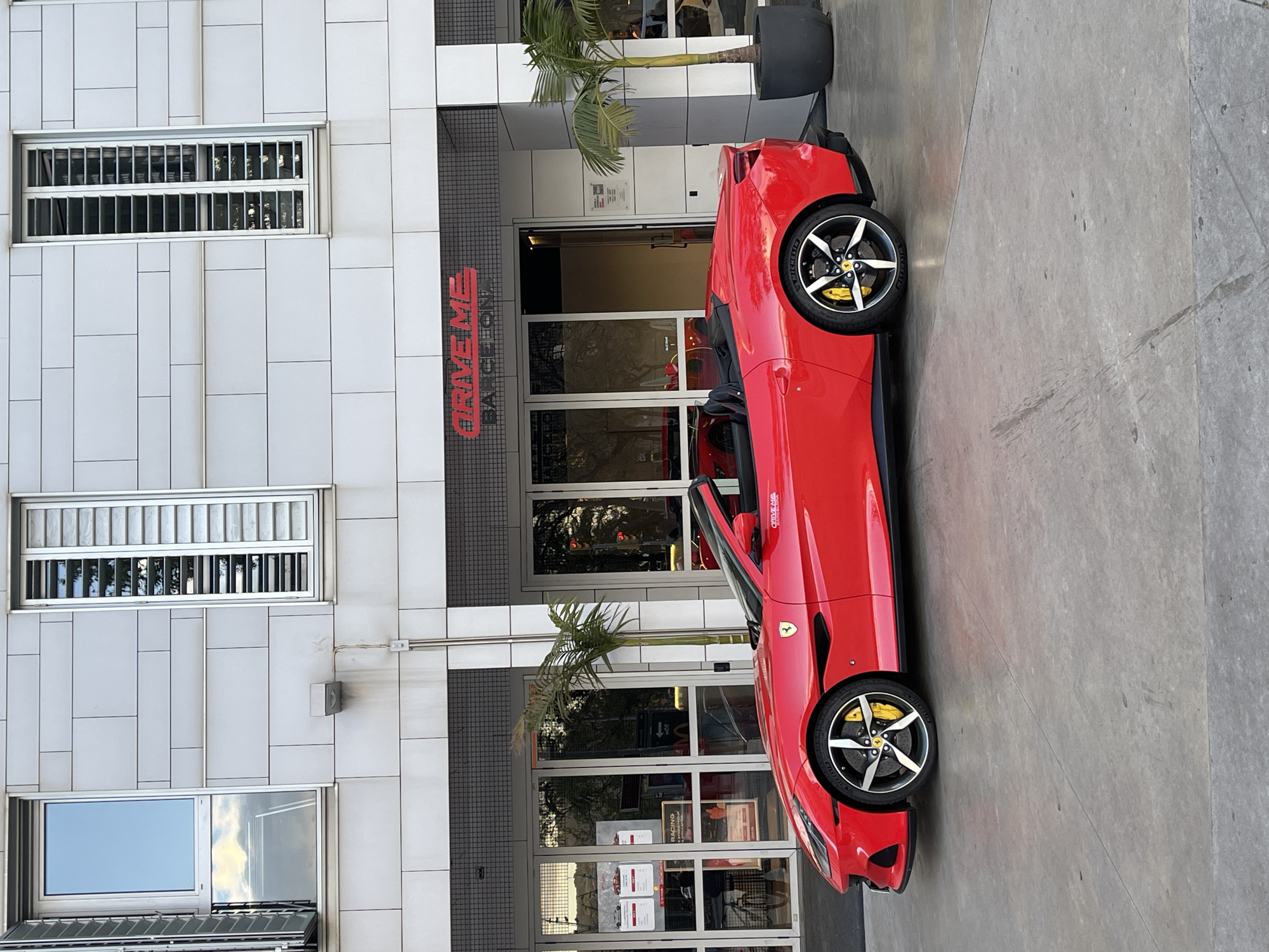 The image shows a red Ferrari parked in front of a building with the sign "DRIVE ME BARCELONA" displayed. The building has a modern facade with windows and a palm tree planted in a pot. The scene appears to be located in Barcelona, Spain, as indicated by the sign. There are no people visible in the image. The car is parked on a concrete surface.
