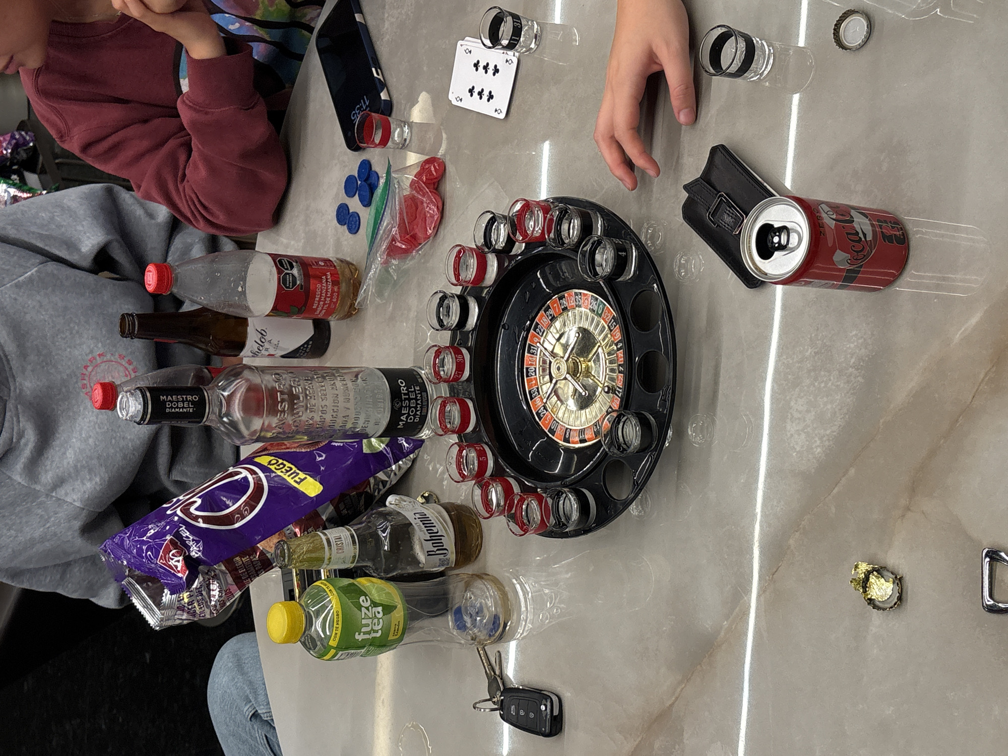 The scene depicts an indoor casual gathering taking place on a light-colored, reflective table, located in San Bernardino Tlaxcalancingo, Mexico. Two individuals are partially visible: one on the left wearing a grey hooded sweatshirt and light blue jeans, and another on the right in a maroon long-sleeved shirt, with their right hand resting on the table.
Numerous objects are arranged on the table. In the upper left, an open bag of "Takis Fuego" chips rests next to a clear glass bottle of "Maestro Dobel Diamante" tequila. To its right, a brown glass bottle of "Chelada" beer stands beside a plastic bottle of "REFRESCO SABOR MANZANA" (apple-flavored soda) with a red cap. A green "Fuze tea" bottle with a yellow cap and a partially consumed "Bohemia Cristal" beer bottle are situated on the left.
The center of the table features a black roulette drinking game set with a golden wheel. Surrounding it are numerous shot glasses; some have red bands, others are clear or have black bands, and several display numbers such as "5," "4," "27 36," and "11 31." To the right of the roulette game, a small red plastic bag containing red candies or similar items is visible, along with five blue circular chips. A stack of playing cards, with an 8 of clubs on top, is positioned further right.
Personal items include a set of car keys (remote and two traditional keys) on the far left, and a black smartphone displaying "11:35" face-down on the right side of the table. A black wallet or card holder with a buckle closure lies beneath an open red "Coca-Cola Zero" can. A crumpled gold foil item and a silver bottle cap are also on the table. The arrangement of objects suggests active engagement in a social gathering involving beverages and games.