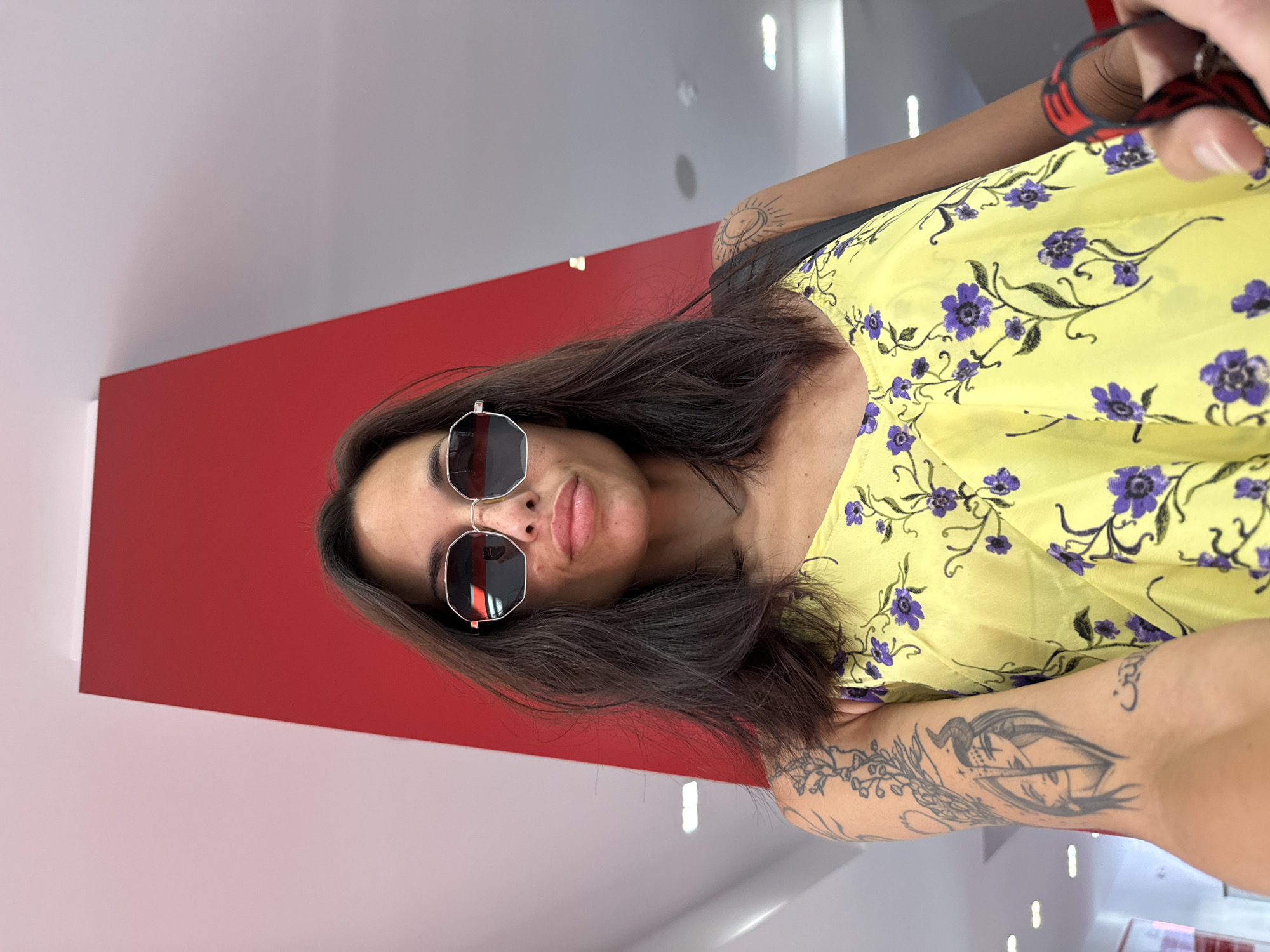 A medium shot captures a woman with dark hair, wearing large, angular sunglasses and a yellow floral print dress. Her left arm, adorned with several tattoos, is extended towards the camera. A red, angular structure dominates the background. The scene is illuminated with bright, artificial lighting. There is no discernible indication of the location being Pleven, Bulgaria.