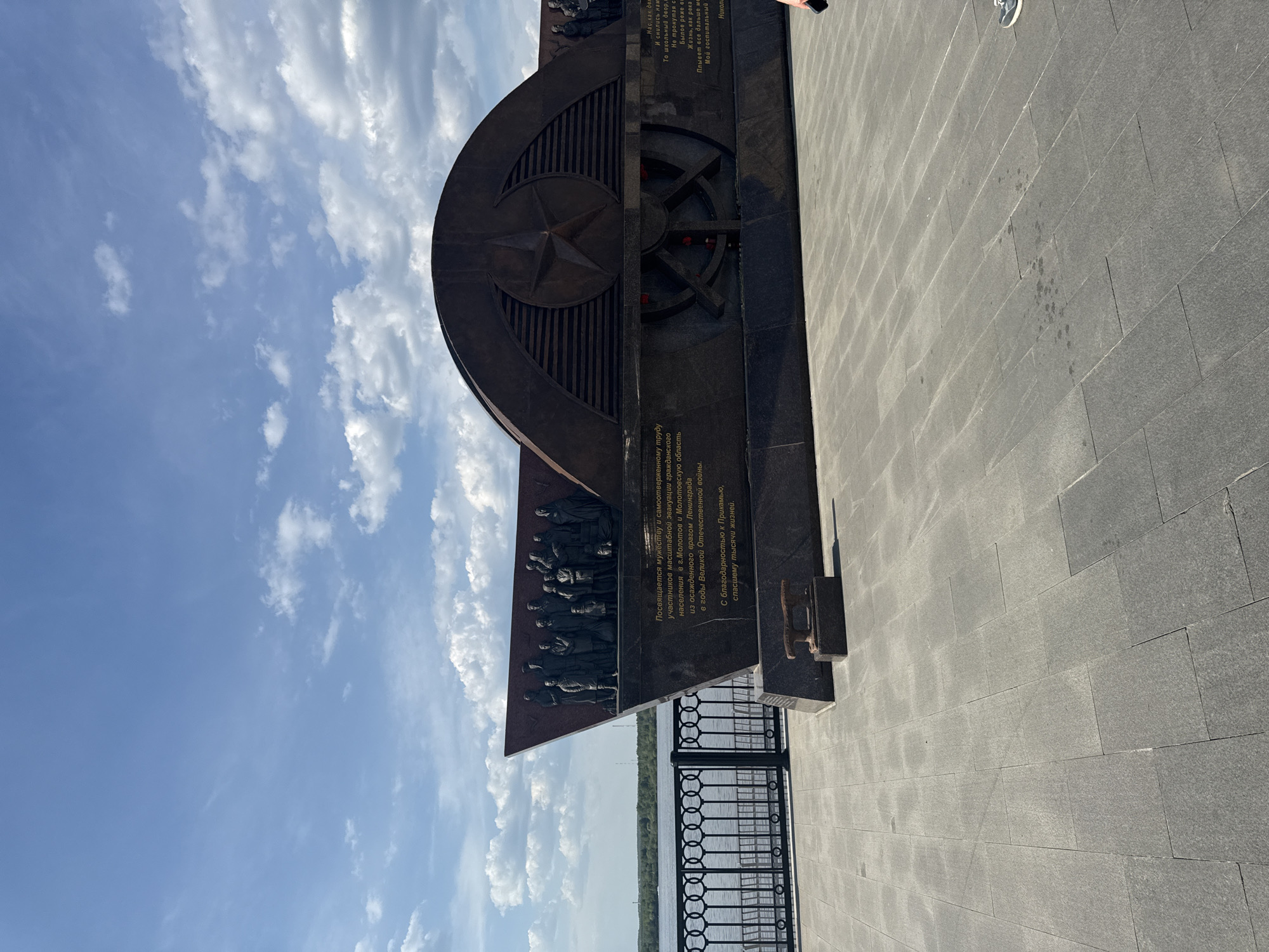 This photograph depicts a monument in Perm, Russia, dedicated to the evacuation of citizens from Leningrad during World War II. The monument is made of dark stone and features a Soviet star and reliefs of figures, likely representing evacuees. Inscriptions in Russian are visible on the monument, expressing dedication and gratitude. The monument is situated on a paved surface near a black metal railing, beyond which appears a view of a body of water. The sky above is blue with scattered white clouds.
