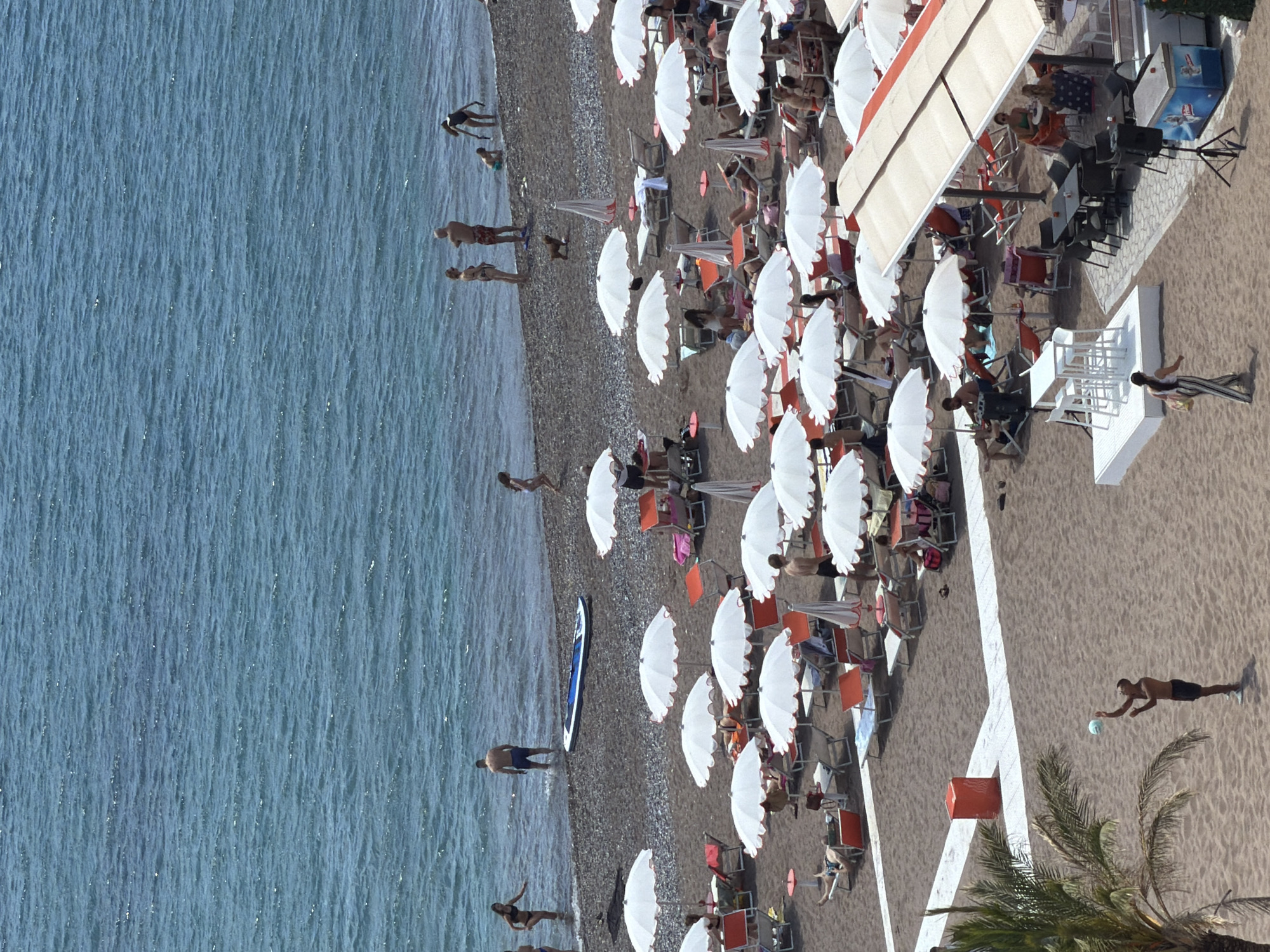 The image shows an aerial view of a beach in Boreti, Montenegro. The scene is composed of a large body of water on the left side of the frame, which appears to be the sea. A narrow strip of pebble beach runs adjacent to the water, where several people are standing or swimming.
The main part of the beach is occupied by numerous white umbrellas, arranged in rows, and accompanied by orange-colored lounge chairs. People are seated under the umbrellas, suggesting a leisure activity. To the right, there is a structure with an awning, implying the presence of a cafe or beach bar. Tables and chairs are visible underneath the awning. A man is playing on the beach next to a palm tree.