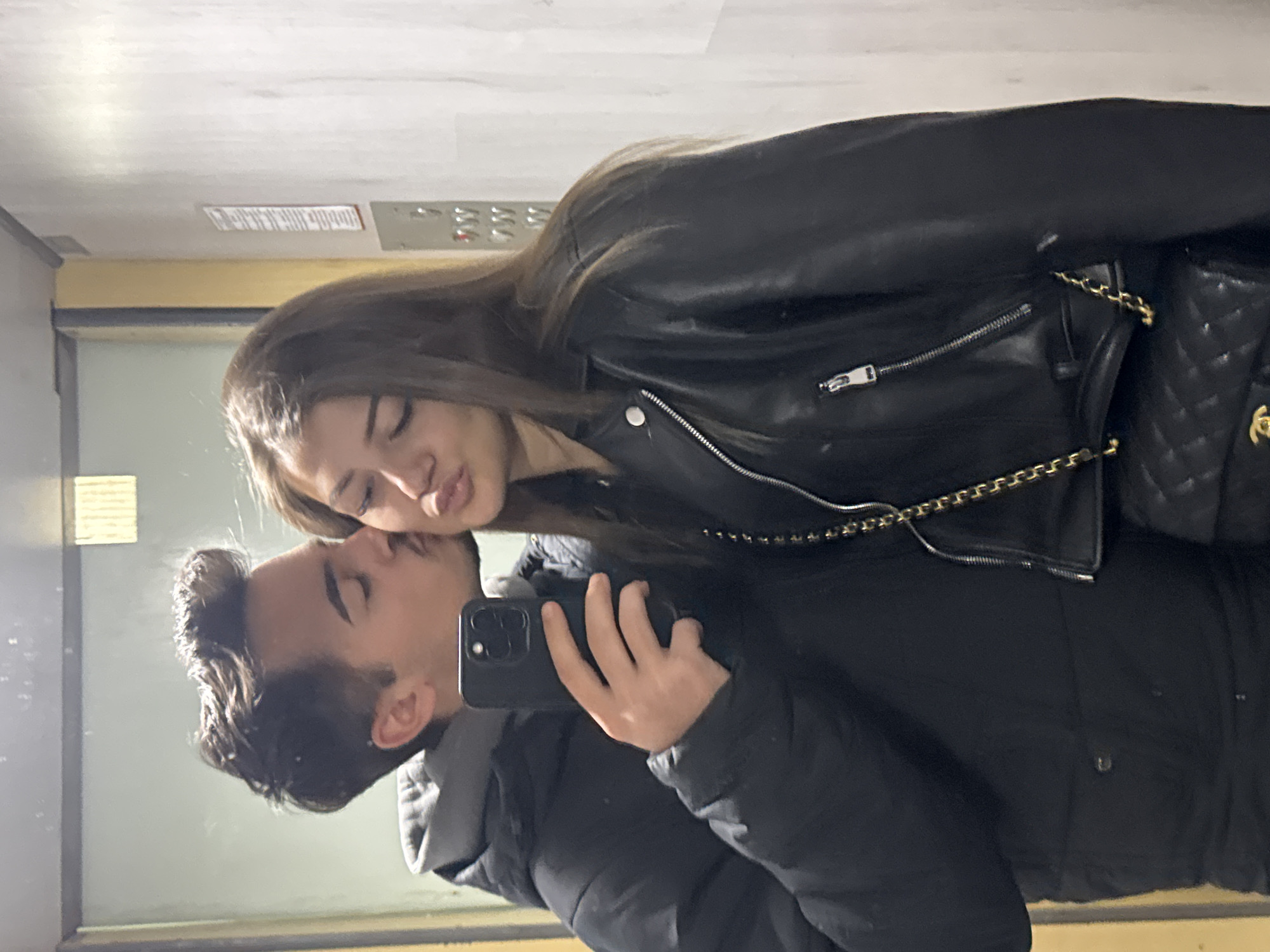 A couple poses for a selfie in an elevator. The woman, with long brown hair, wears a black leather jacket and a matching purse with a gold chain. She leans towards the man, puckering her lips for a kiss. The man, holding a black smartphone to take the picture, wears a dark puffer jacket with a grey hood. The elevator interior is utilitarian with metal panels and control buttons visible in the background. The location is likely in Sofia, Bulgaria, but there is no verifiable information to confirm this, the location is only the user's inference.