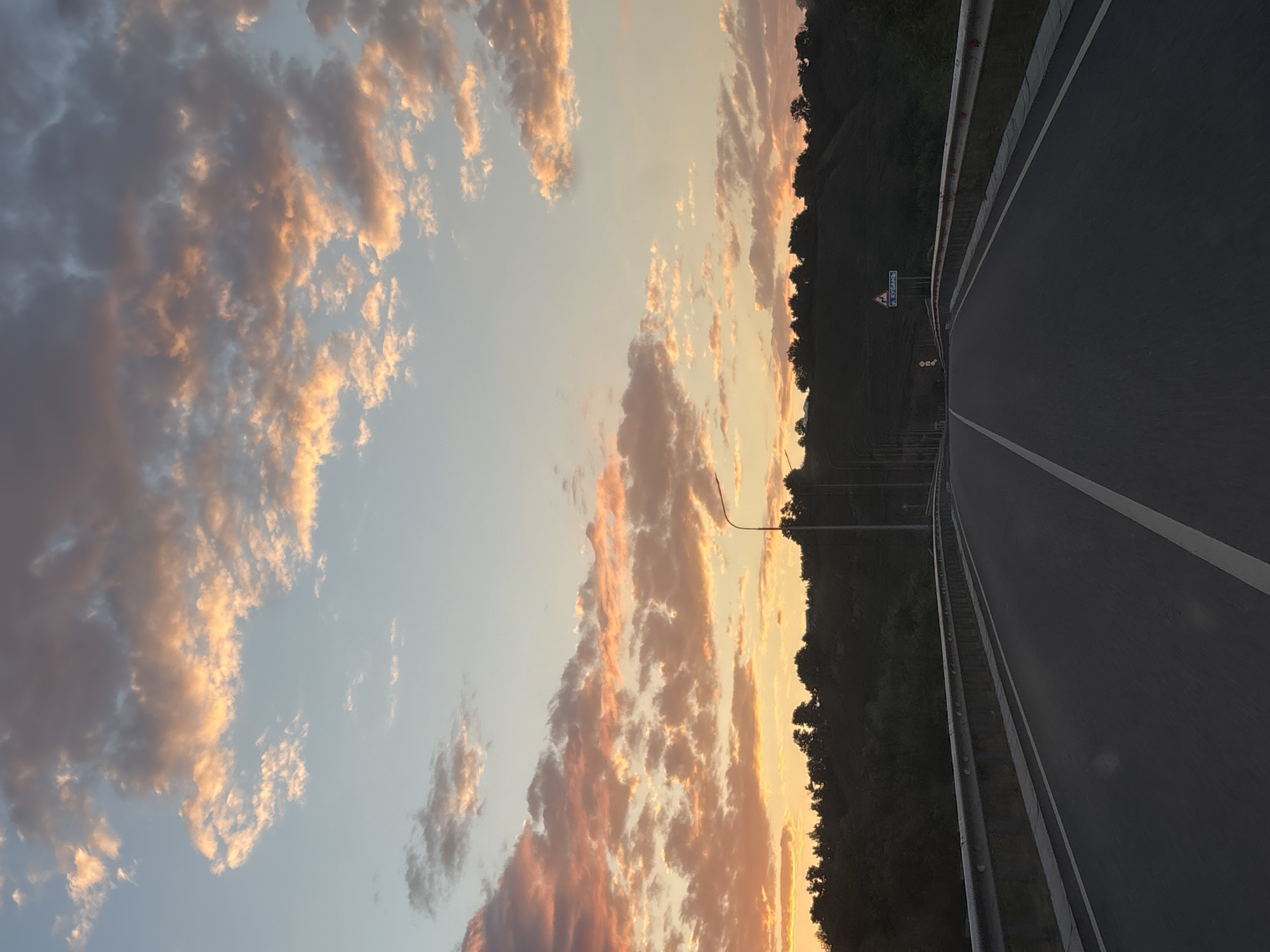 This is a photograph of a road leading through a rural landscape at dusk. The sky is filled with a vibrant sunset, with clouds illuminated in shades of orange, pink, and yellow. A dark silhouette of trees and a rolling hill forms the horizon. The road itself is asphalt with a white line marking the center. A metal guardrail runs along the side of the road. Streetlights are visible in the distance. A triangular road sign with Cyrillic text is posted on the right side of the road. The image does not contain any people.