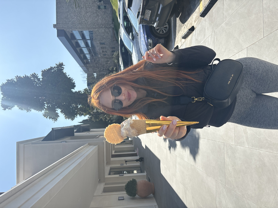 A female subject with long, reddish-brown hair, dark sunglasses, a black padded jacket, grey leggings, and a black cross-body bag stands outdoors, extending an ice cream cone toward the viewer. Her left hand, featuring light-colored manicured nails, grips the gold-wrapped cone, which is topped with multiple scoops of ice cream and a circular waffle biscuit. The scene is set in Budva, Montenegro, under clear daylight conditions. To the subject's left, a light-colored, multi-story building with dark awnings fronts a grey tiled pedestrian area, where a large terracotta pot with greenery and a black kick scooter are visible. A large, densely foliated tree is positioned centrally in the background against a bright blue sky. To the right, a parking area contains several parked vehicles, including a dark SUV displaying the rear license plate "BG 2705 CE". Further behind the parking area, a building with a stone-like facade and dark windows is present.