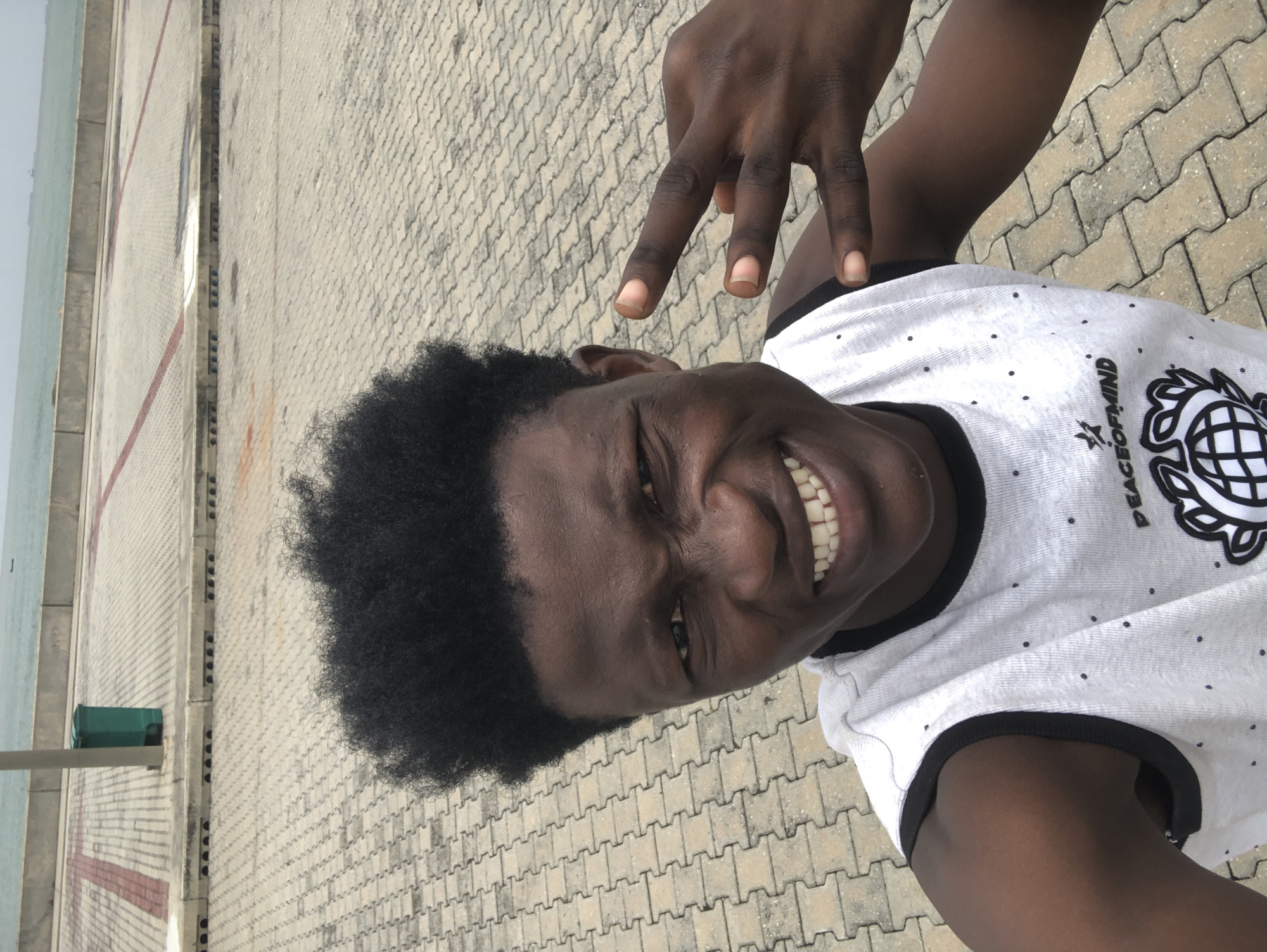 A young Black male with dark, curly hair smiles and makes a peace sign with his right hand. He is wearing a white, sleeveless shirt with black trim and a pattern of small black dots. The shirt also has a graphic with text that reads "PEACEOFMIN" and a circular emblem. He is outdoors on a paved area with interlocking brick pavers. In the background, there is a concrete barrier and beyond that, a body of water, likely the ocean. A green trash can and a lamppost are visible to the left. The scene appears to be in a public outdoor space, potentially along the coast in Ikoyi, Nigeria.
