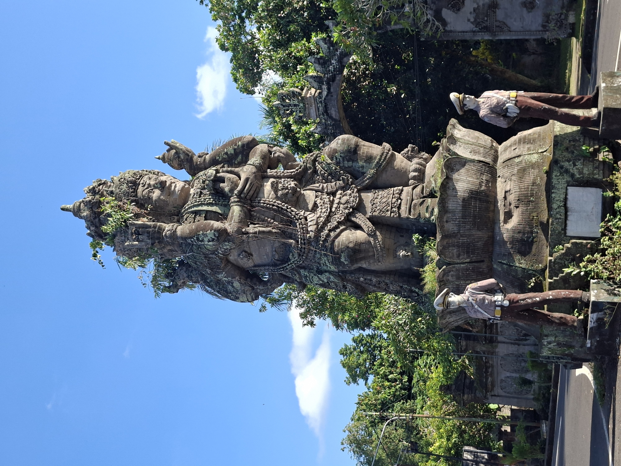 The image captures a monumental stone statue of a multi-armed deity, weathered and partially covered in moss, standing prominently in Banjarangkan, Indonesia. The statue is positioned on a lotus-shaped pedestal, with two life-sized statues of what appear to be police officers flanking it, one on each side. These figures are dressed in traditional police uniforms, complete with hats, shirts, belts, and trousers, and are positioned with their hands behind their backs. The background features lush green trees and a clear blue sky with scattered clouds. An arched gateway, also made of stone and adorned with traditional carvings, is visible behind the main statue. The scene is set outdoors, likely in a public space or at the entrance to a significant location, with a road visible in the foreground.