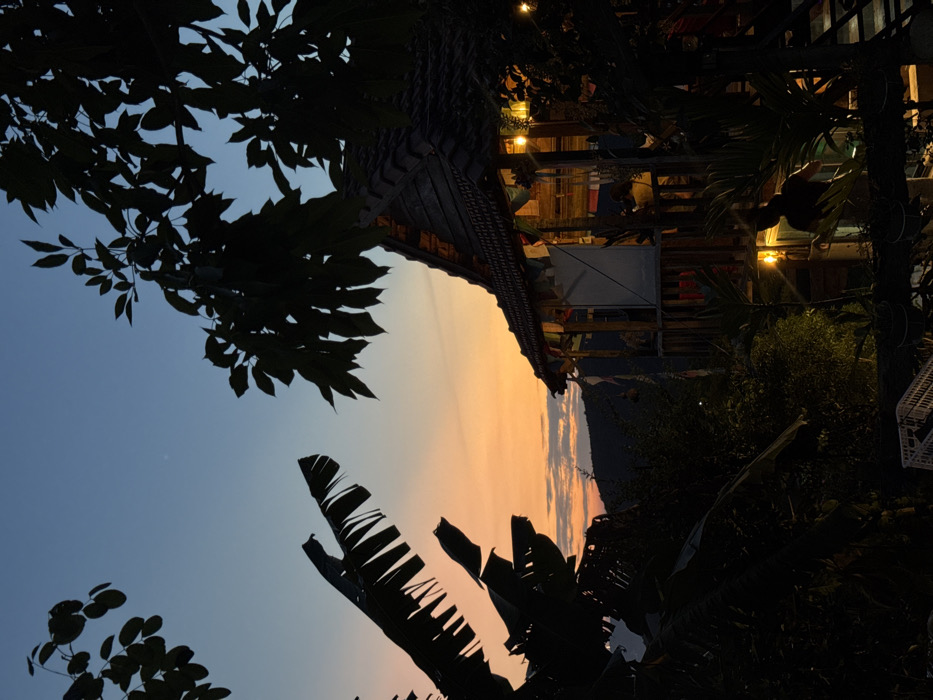 The image depicts a scene at dusk in Nha Trang, Vietnam, featuring a multi-story wooden structure, likely a cafe or guesthouse, nestled amidst dense tropical foliage. The sky transitions from a deep blue at the top to vibrant orange and yellow hues near the horizon, with a faint line of distant hills silhouetted against the colorful light.
The central subject is a building with a steeply pitched, dark tiled roof. Its upper level features an open wooden balcony adorned with multiple colorful banners or prayer flags strung along the railing. String lights illuminate the exterior and interior spaces of the building. On the upper balcony, one person is visible, seated and mostly silhouetted against the bright interior. A second individual is partially visible on the lower level, also silhouetted against an interior light source, appearing to stand or move within the space.
The foreground and left side of the image are dominated by the dark silhouettes of large banana leaves and other broad-leafed trees, framing the scene. Additional dense vegetation is visible below and around the structure, further integrating it into the natural environment. The overall lighting suggests the period immediately following sunset.