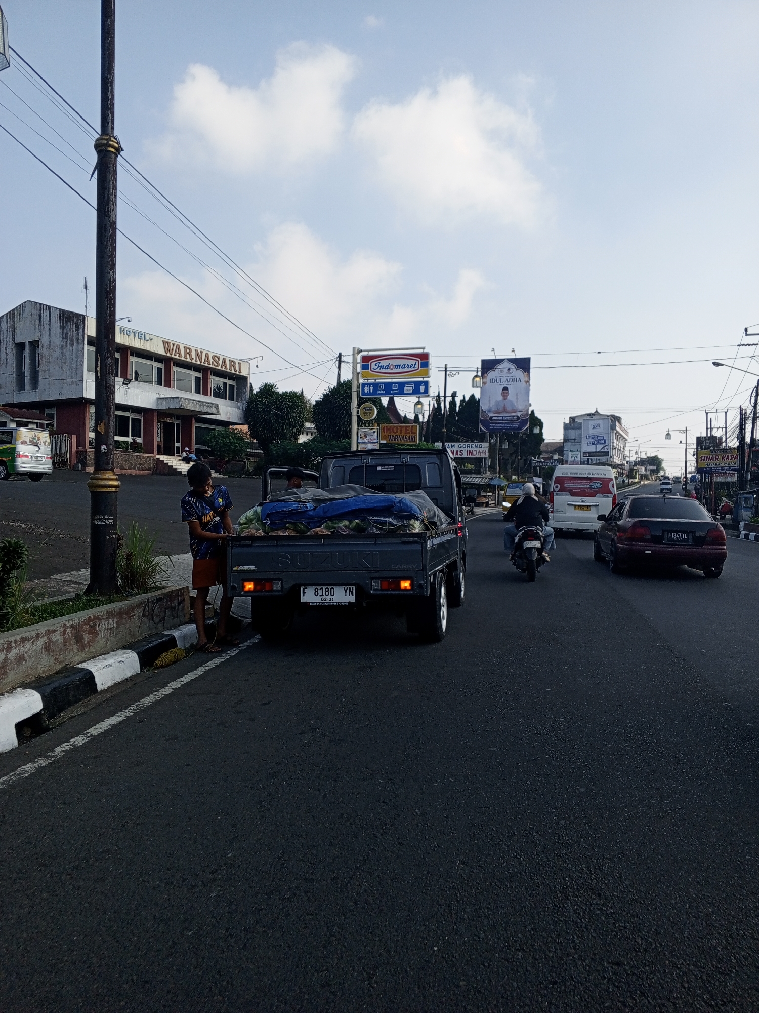 The image captures a bustling daytime street scene in a city, identifiable by the presence of an Indomaret convenience store sign and Indonesian license plate formats on visible vehicles. On the left, a multi-story building with "HOTEL WARNASARI" prominently displayed on its facade stands alongside the road. In the foreground, a tall, dark utility pole with overhead wires extends upwards.
A dark gray Suzuki Carry pickup truck is parked or stopped on the left side of the road, close to a curb painted in black and white stripes. Its tailgate is open, revealing a cargo bed filled with numerous items, possibly produce like large fruits or vegetables, partially covered by a blue tarp. A young male in a blue patterned shirt and brown shorts stands next to the open tailgate, appearing to tend to the cargo. He is wearing flip-flops. Another person is visible inside the cab of the pickup. The truck's license plate, "F 8180 YN," is clearly visible. A coil of yellow material rests on the curb near the young man's feet.
Further down the road, traffic is moving. A motorcyclist in a white helmet and dark jacket is riding on the asphalt road, positioned behind the pickup truck. A white passenger van with "Rhis.id" printed on its side is visible further ahead, followed by a black sedan with the license plate "F 1347 YL." In the background, several billboards are visible; one prominently features "IDUL ADHA" with an image of a man, and another advertises "SINAR KAPA." The sky above is clear blue with scattered white clouds. The overall environment suggests an urban or semi-urban commercial area with various businesses lining the street.