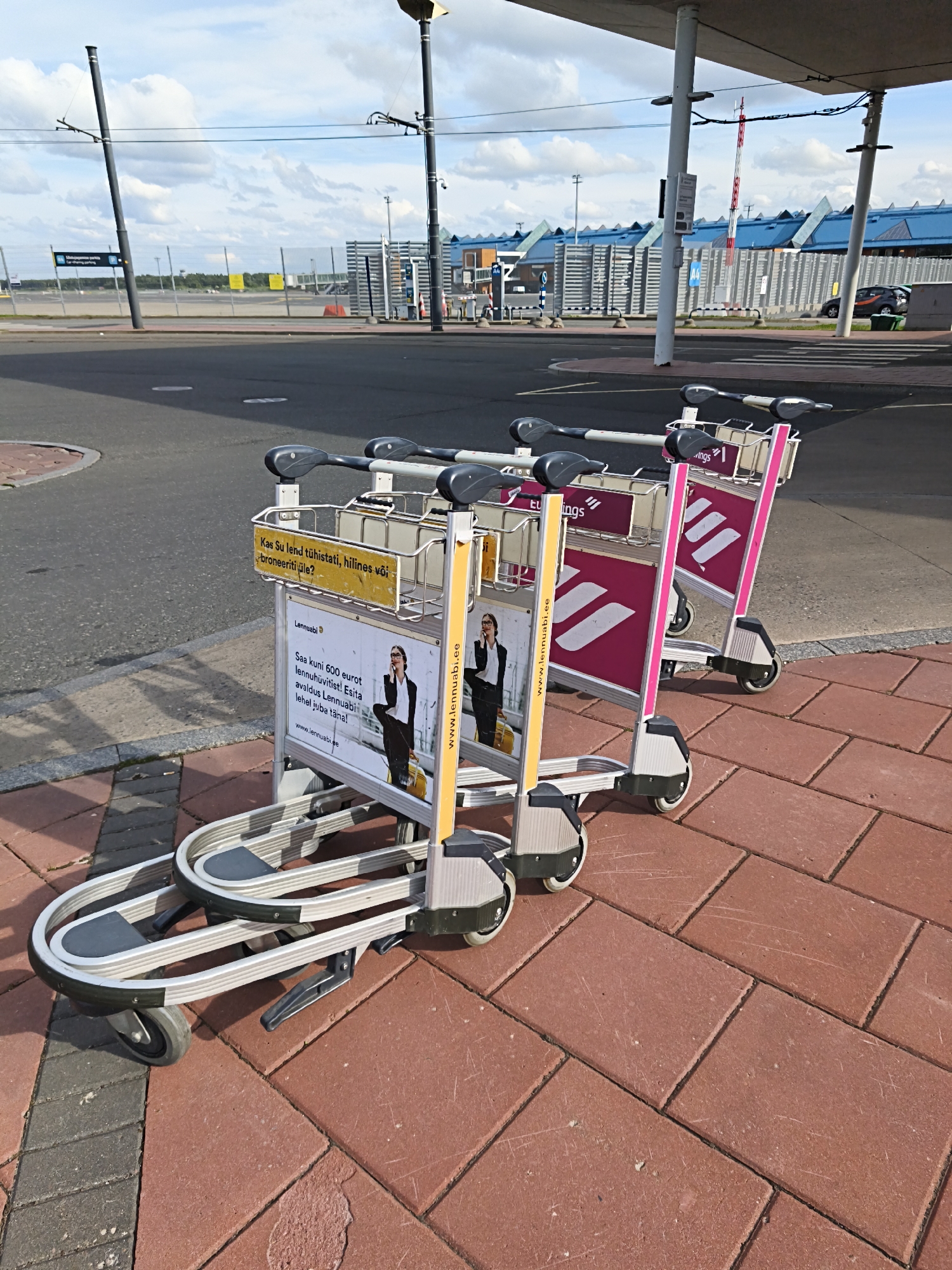 The image shows several luggage carts parked on a red brick sidewalk in front of an airport terminal. The carts have various advertisements on them, including one for "Lennuabi" and another with text in Estonian. In the background, there is a road, fencing, and the modern architecture of the airport building. The sky is partly cloudy. This scene is likely at Tallinn Airport (TLL) in Tallinn, Estonia, given the Estonian language on the advertisements.