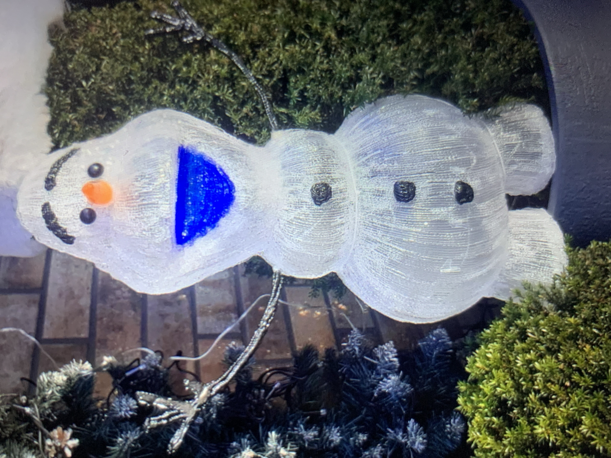 The image shows a snowman decoration, resembling Olaf from the movie Frozen, placed outdoors. The snowman is made of white, sparkly material and has blue lights for its mouth. It has black eyes and buttons, and an orange nose. The snowman is positioned in front of a brick wall, with a Christmas tree and green shrubbery to its sides. The overall scene suggests a festive outdoor decoration, likely for the winter holiday season. There is no information in the image to suggest a location in Lymanka, Ukraine, or any other specific city.