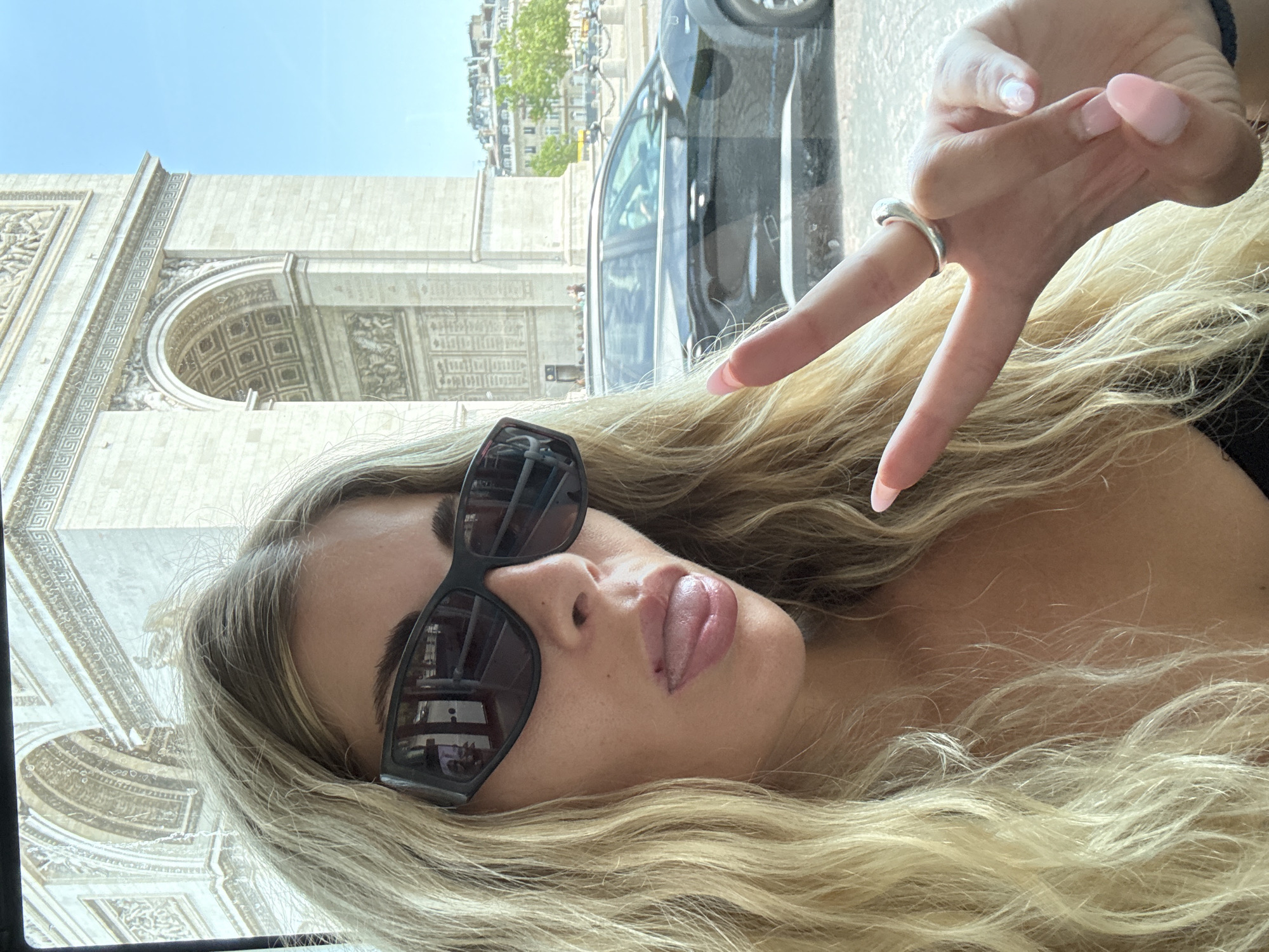 A woman with long blonde hair, wearing large black sunglasses and a silver ring on her right index finger, sticks her tongue out at the camera while making a peace sign with her right hand. She is positioned in the foreground, with the Arc de Triomphe visible in the background in Paris, France. A black car is driving by in the middle ground, and a bright blue sky is visible above. The image appears to be taken from inside a vehicle.