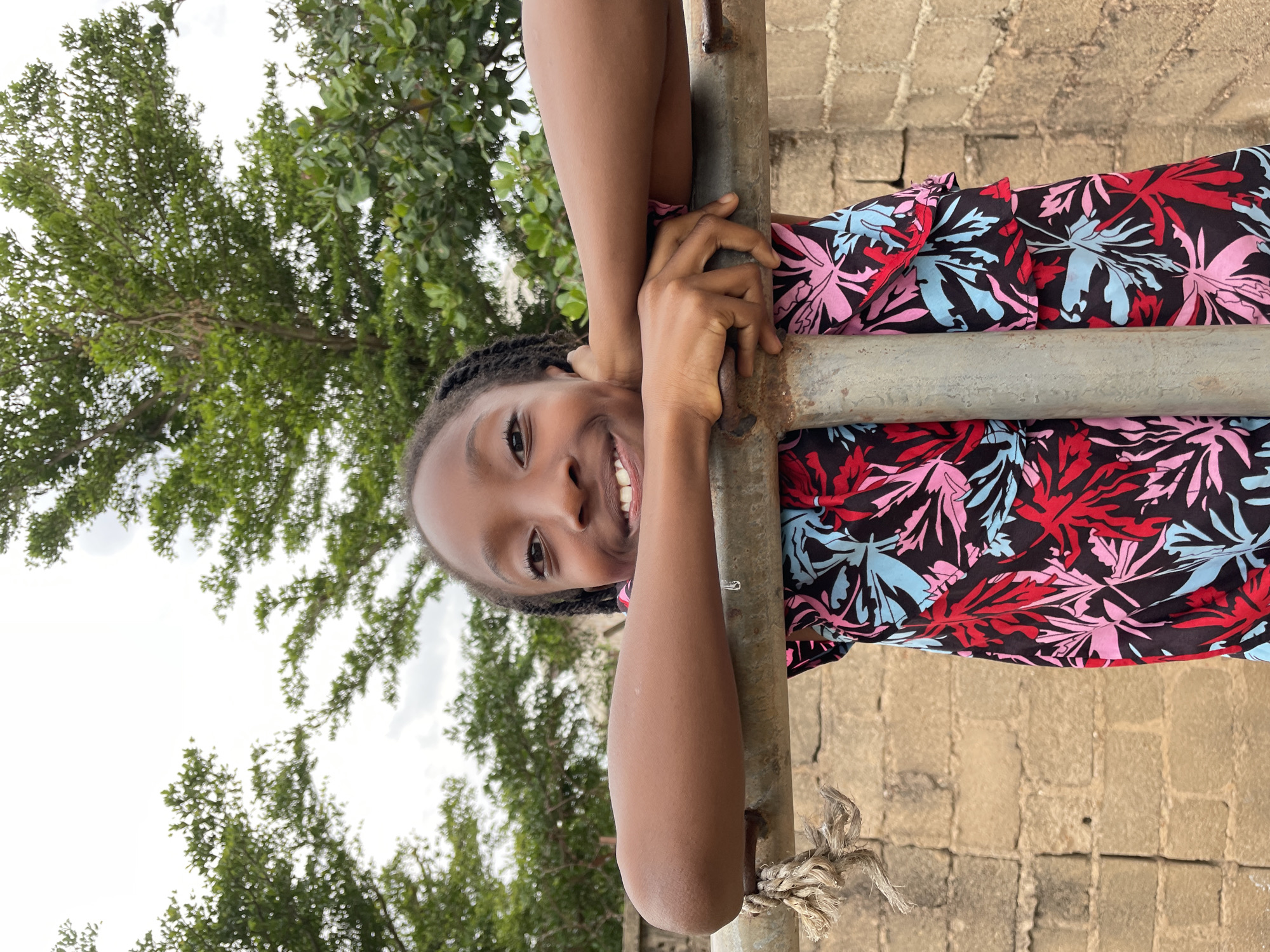 A young Black girl with braided hair smiles broadly while leaning on a metal railing in an outdoor setting. She is wearing a black dress with a vibrant floral print of red, pink, and light blue leaves. Her arms are crossed over the railing, with her hands resting on it. Behind her, a brick wall is visible, and beyond that, lush green trees under a cloudy sky. The scene appears to be a public space, possibly a park or a schoolyard, in Jos, Nigeria. The lighting suggests it is daytime.
