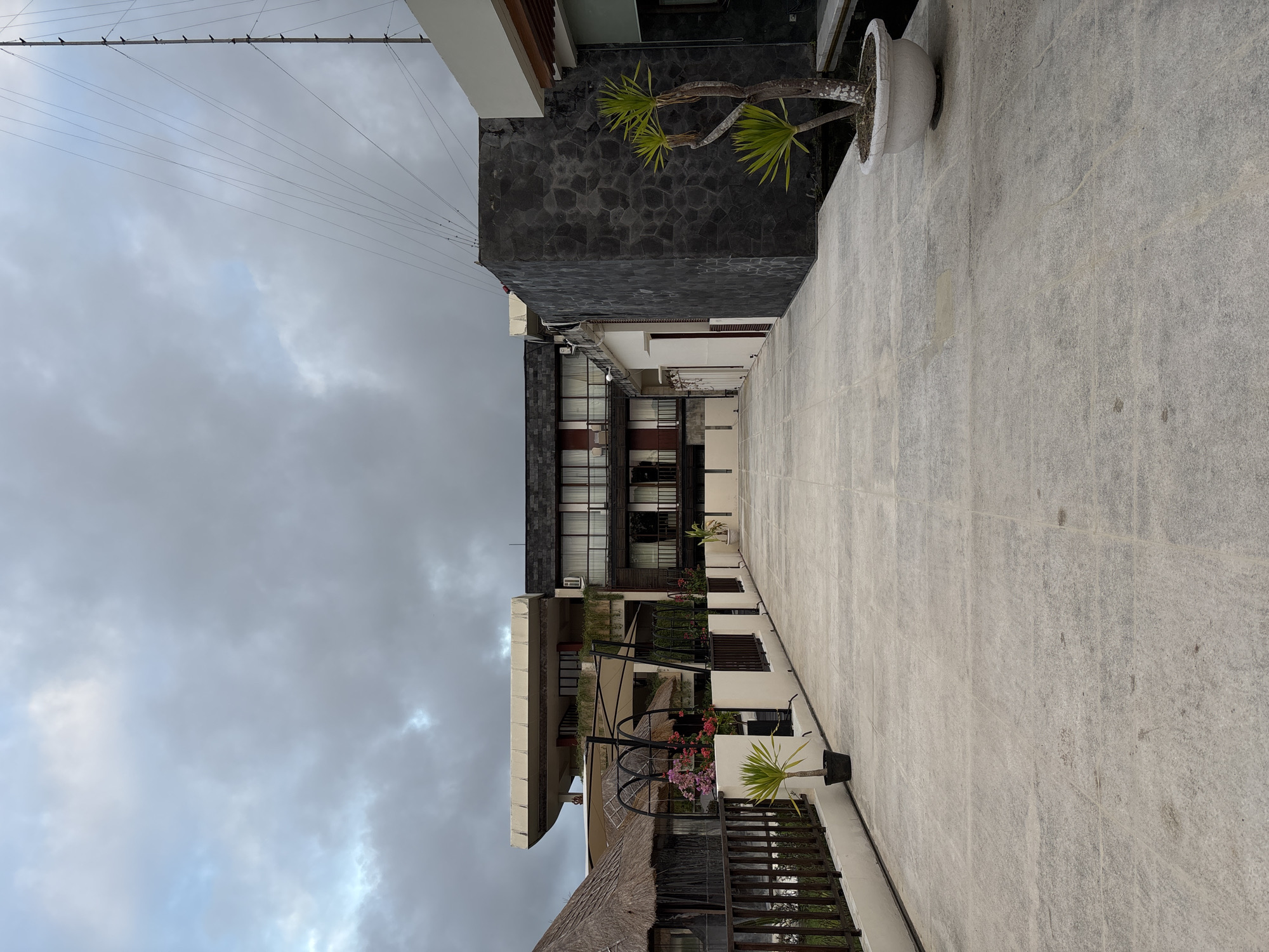 The image depicts a rooftop terrace in Kuta Selatan, Indonesia, under a cloudy sky. The terrace is paved with large, light-colored tiles and features a dark stone wall on the right side, with a potted plant in front of it. To the left, a dark wooden railing separates the terrace from a lower level with thatched-roof structures and greenery. In the background, a multi-story building with dark wood accents and balconies is visible, along with another building with a more modern facade. A tall antenna structure with numerous wires extends upwards into the sky on the far right. No people are present in the scene.