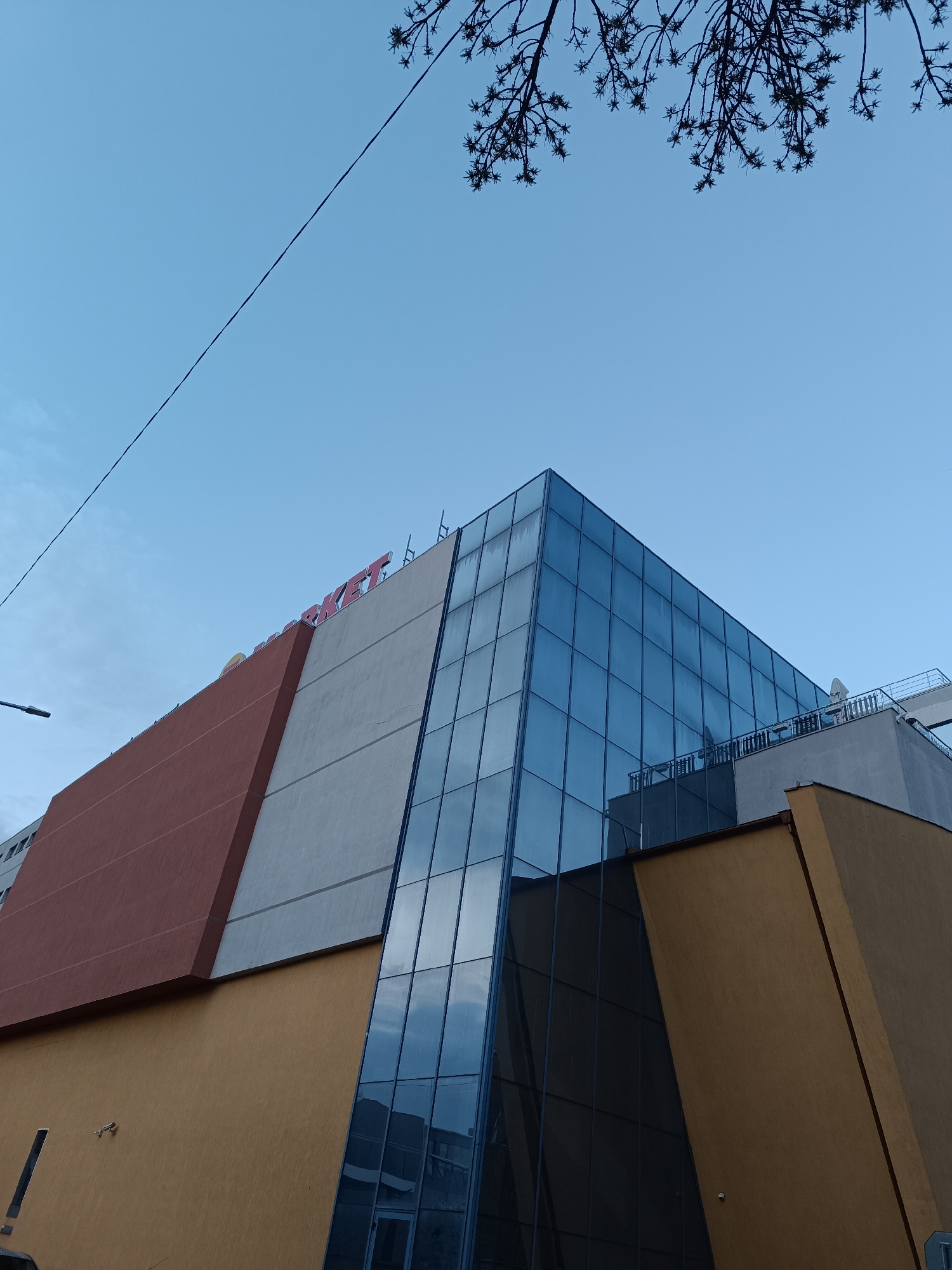 A low-angle, daytime shot of a modern building, likely a commercial property, in Pleven, Bulgaria. The building features a combination of materials, including a glass facade with a grid-like pattern, light gray concrete, and brown stucco. The facade also shows a section of light-brown stucco. The building is multi-storied.
A red sign with the word "MARKET" is visible on top of the building. The sky is blue, with a few branches visible in the upper-right corner. Power lines are visible against the sky in the upper-left corner.