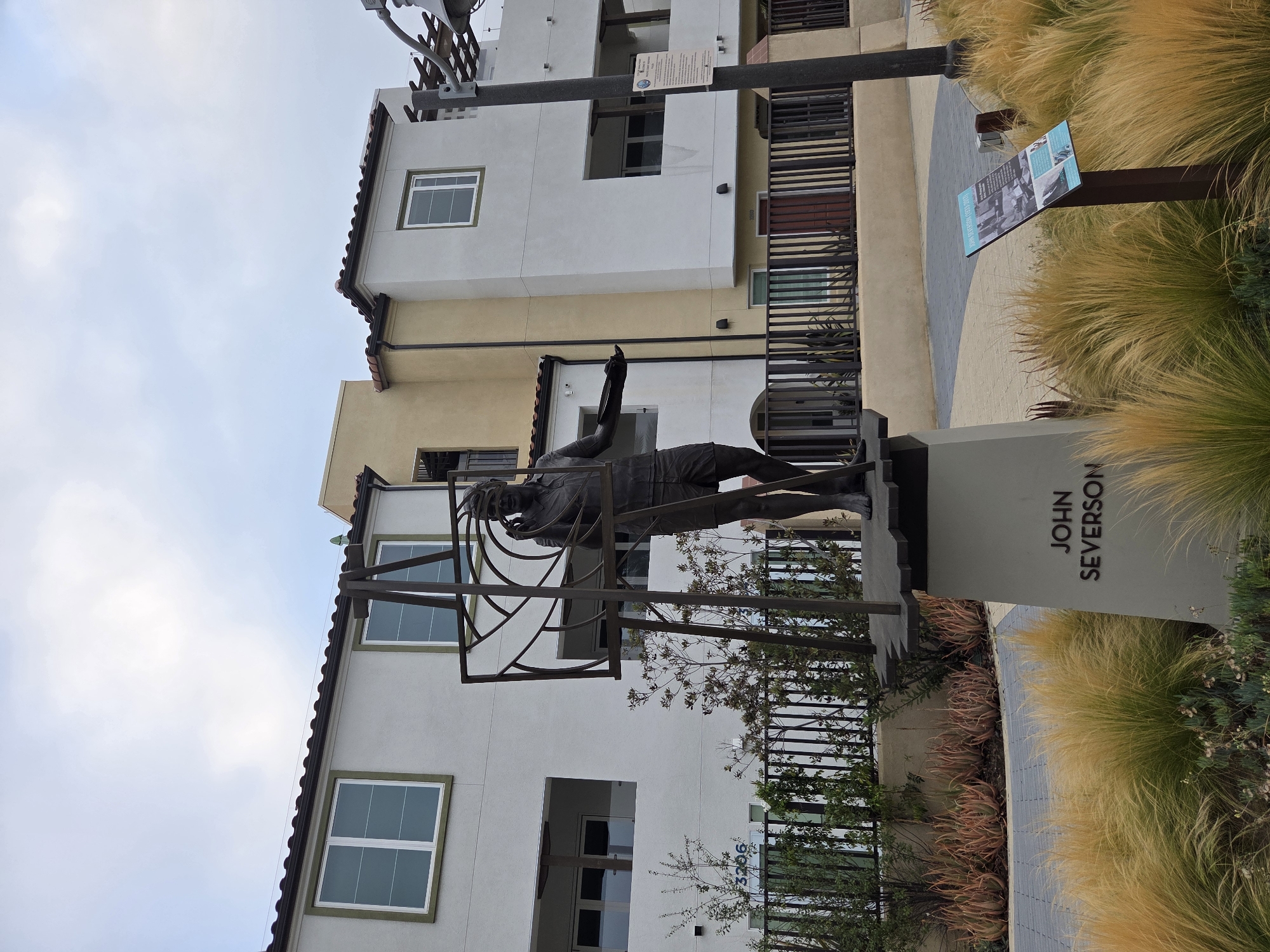 The image shows a statue of a man, likely John Severson, in Dana Point, California. The statue depicts the man in a dynamic pose, with one arm raised as if reaching for something. He is dressed in athletic attire, with shorts and a shirt. The statue is made of dark metal and is placed on a pedestal that has "JOHN SEVERSON" inscribed on it.
The setting is an outdoor urban environment, possibly a park or a public plaza. There are modern buildings in the background, featuring white and yellow facades and windows. A metal fence is visible, separating the area with the statue from the buildings. In the foreground and to the right, there are tall, golden-colored ornamental grasses and some succulents. A sign with text and images is also visible near the grasses.
The sky is overcast, suggesting it might be a cloudy day, or perhaps early morning or late afternoon. The overall mood of the image is serene and contemplative, focusing on the statue as a tribute. The urban architecture contrasts with the natural elements of the plants. The inscription on the pedestal clearly identifies the subject of the statue.