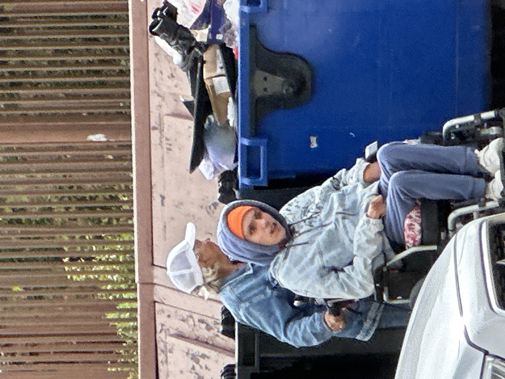 Two individuals are situated near large blue dumpsters. In the foreground, a person in a wheelchair is wearing a light blue hooded sweatshirt and an orange beanie. They are seated with blue sweatpants and white sneakers. Behind the person in the wheelchair, another individual, partially visible, is wearing a denim jacket and a white baseball cap. They appear to be pushing the wheelchair. The environment suggests an outdoor urban setting with a chain-link fence in the background and a silver vehicle in the lower left corner. The overall scene depicts a moment of assistance or transit. The image does not contain any elements that indicate the location in Podolsk, Russia.