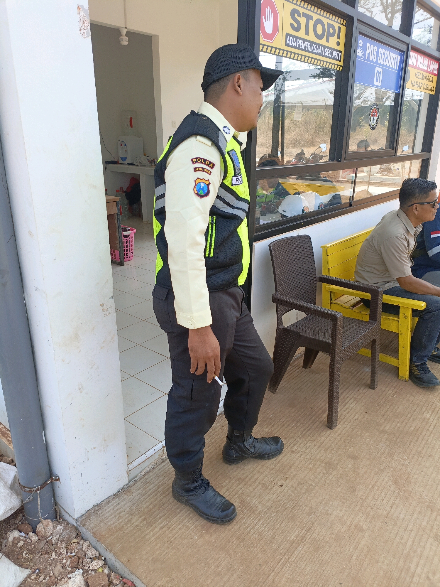 An Indonesian security guard, wearing a black cap, a tan long-sleeved shirt with a yellow and black vest, and black pants and boots, stands smoking a cigarette to the left of the frame. He is positioned near a white wall and a gray pipe. Behind him, partially visible through a doorway, is a room with a water dispenser and some shelves. To the right of the guard, a windowed booth is visible with various signs. One sign reads "STOP! ADA PEMERIKSAAN SECURITY," another reads "POS SECURITY 01," and a third says "AWAI WAJIB LAPOR HELM KACA HARAP DIBUKA." Inside the booth, several motorcycle helmets are visible. To the right of the booth, two men are seated on a yellow bench. One man, wearing a tan shirt and sunglasses, is looking to the right. The other man, wearing a blue jacket with red and white trim, is partially visible. The ground in the foreground is concrete with a textured pattern, and there are some rocks and debris scattered around. The background appears to be an outdoor, dry environment with vegetation.