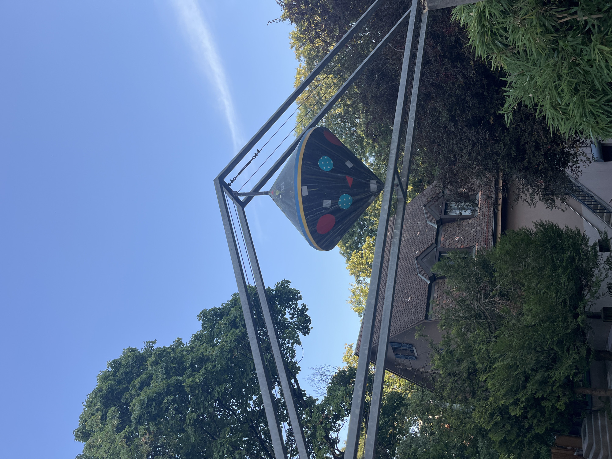 An outdoor, low-angle, eye-level shot shows a conical, dark gray object suspended from a metal structure against a clear blue sky. The object, decorated with colorful circular and square shapes in red, blue, and yellow, hangs from wires attached to the metal frame. Behind the suspended object and metal structure, a brown-tiled roof house with dormer windows is partially visible amidst lush green trees. The overall scene suggests an urban or suburban environment with ample vegetation. The location is the Parc des Bastions in Geneva, Switzerland. There are no people present in the image.