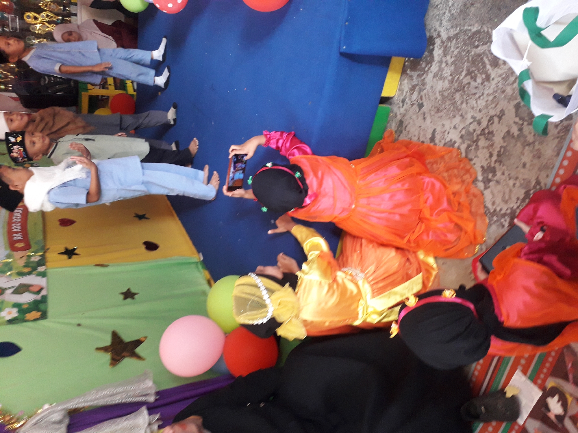 The image depicts an indoor scene, possibly a school or community event in Cankuang, Indonesia. A group of children are dressed in colorful attire. In the foreground, a young child wearing a black hijab and an orange dress is seated, holding a mobile phone and appears to be taking a picture or recording a video. To their left, another child in a black hijab and a bright orange and yellow dress is kneeling. Several other children are visible in the background, some in similar traditional clothing. The environment features colorful backdrops, including green and yellow fabric, as well as balloons. The floor is covered with a blue mat and a patterned rug. In the upper right corner, trophies are visible, suggesting a celebratory occasion.