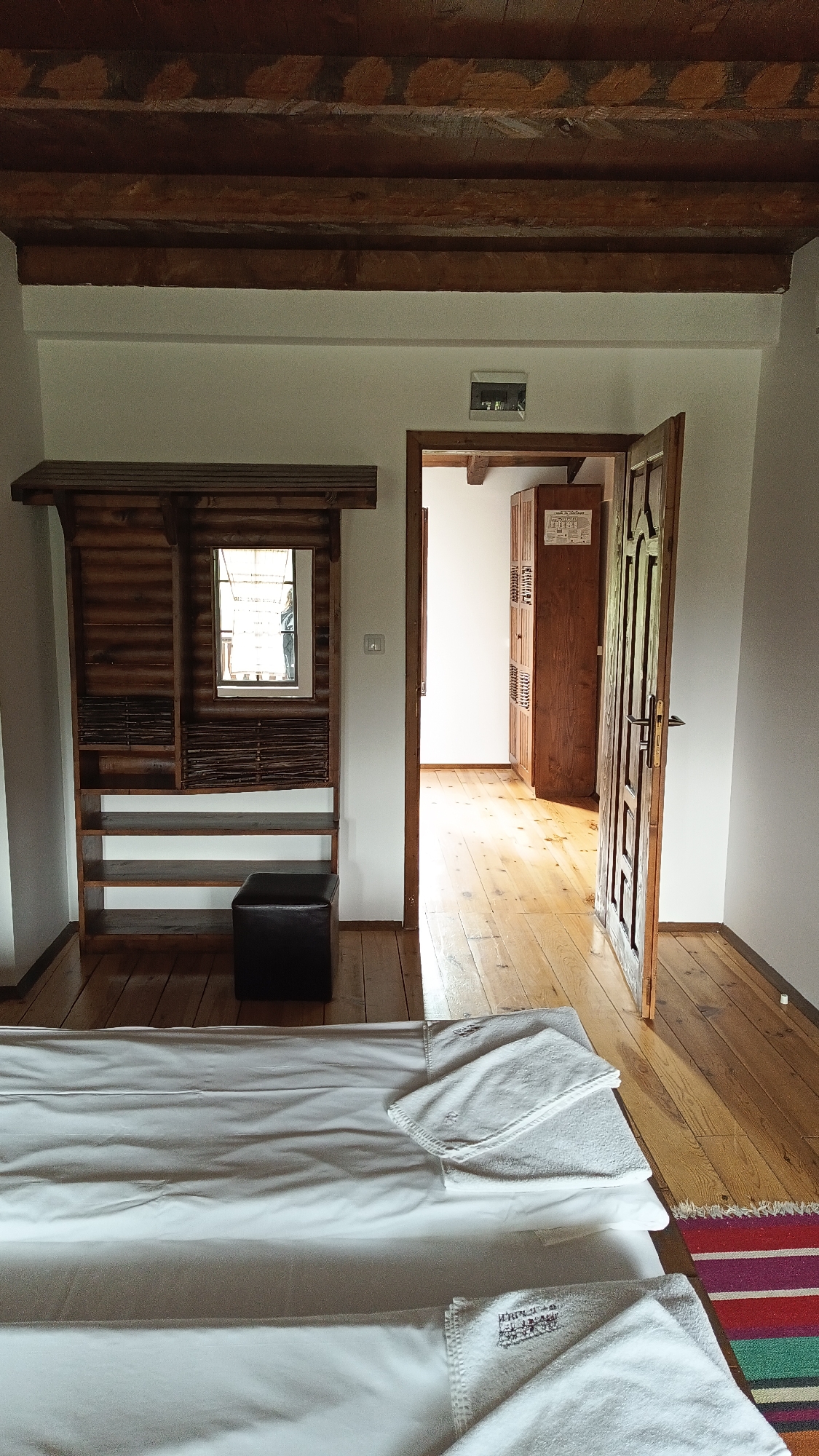 The image shows a bedroom interior in Vrata, Bulgaria. The room features exposed wooden beams on the ceiling and light hardwood flooring. Against the wall on the left is a rustic wooden unit with shelves, a mirror, and woven details. A dark-colored ottoman sits near this unit. In the foreground, a bed is partially visible, covered with white linens and neatly folded white towels. An open wooden door reveals a hallway leading to another room with a wooden cabinet. The walls are painted white. An electrical box is mounted above the doorframe.