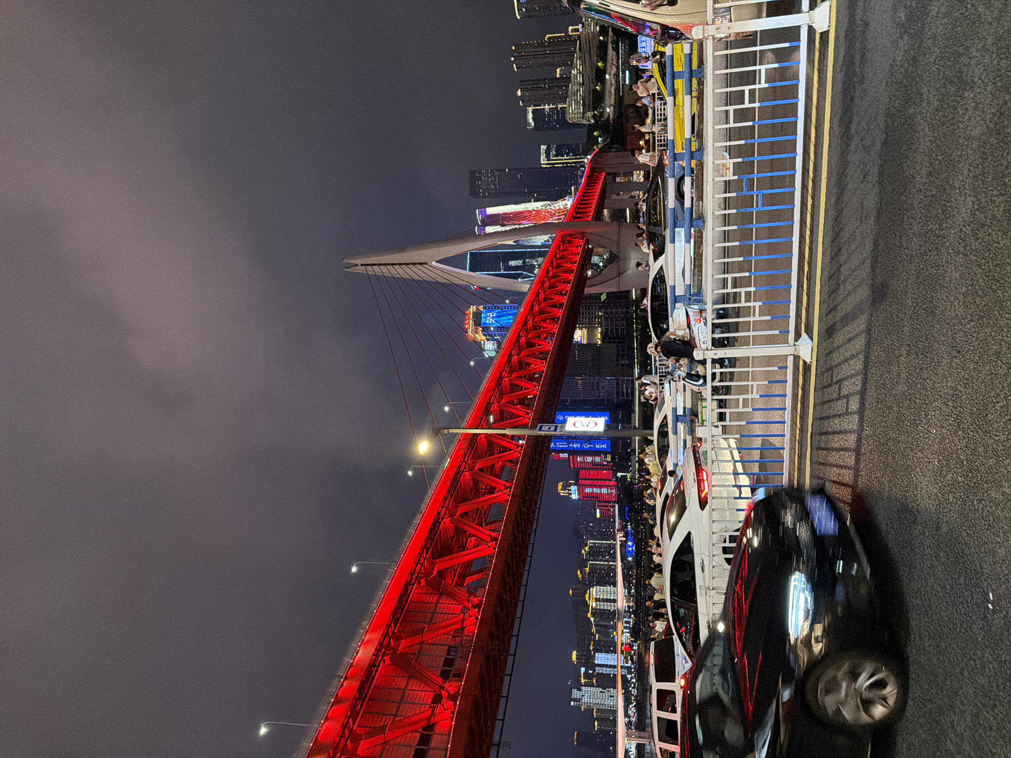 In Yuzhong District, China, a brightly lit red suspension bridge spans a body of water at night. The bridge's red illumination contrasts sharply with the dark, cloudy sky above. In the background, a city skyline is visible, with many buildings illuminated by colorful lights. Traffic is present on the bridge and on the road in the foreground, with cars and a scooter visible. People are standing on a pedestrian walkway on the bridge, observing the city and the bridge itself. A blue and white metal barrier separates the road from the walkway. The overall scene depicts a vibrant urban nighttime environment.