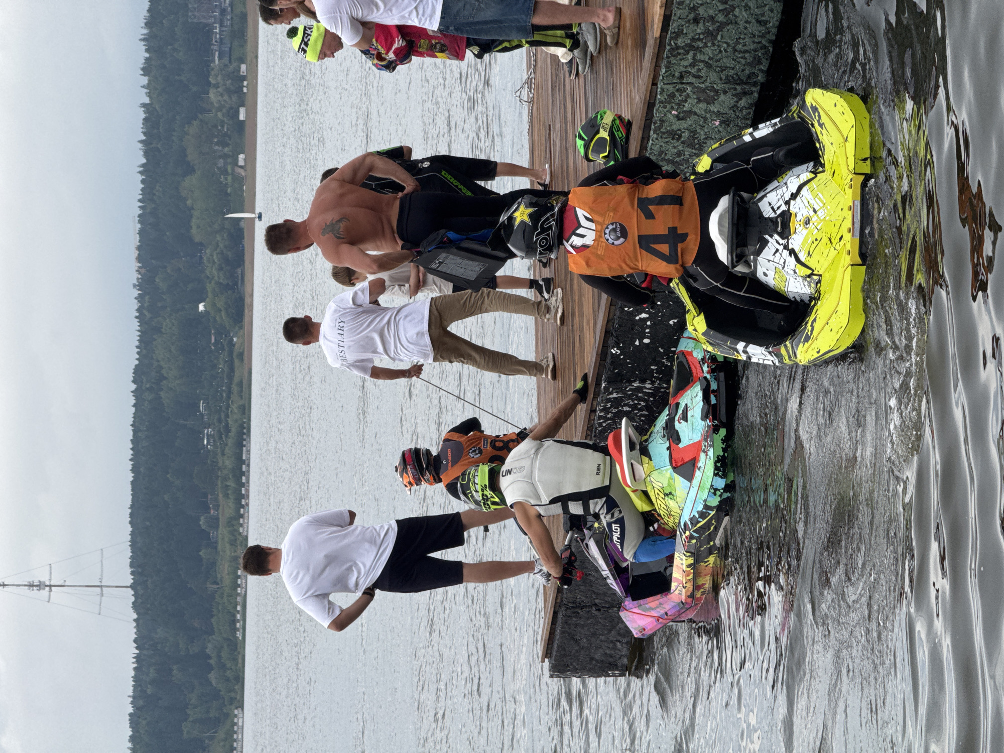 In Izhevsk, Russia, a group of people are gathered on a wooden dock beside a body of water, with a distant forest and radio tower visible under a cloudy sky. Two individuals are seated on brightly colored jet skis, one with a neon yellow and white design and the other with a multi-colored abstract pattern. Both jet skis are partially submerged in the water.
Several other individuals stand on the dock, observing the jet skis. Some are wearing athletic attire, including helmets and life vests, suggesting they are participants in a water sport event. One person, facing away from the camera, displays a tattoo on their back. Another person wears a yellow beanie with green accents.
The scene appears to be set on the shore of the Izhevsk Reservoir, a large artificial lake on the Izh River, commonly used for recreational activities. The overall atmosphere suggests a pre-race or preparation phase for a jet ski event.