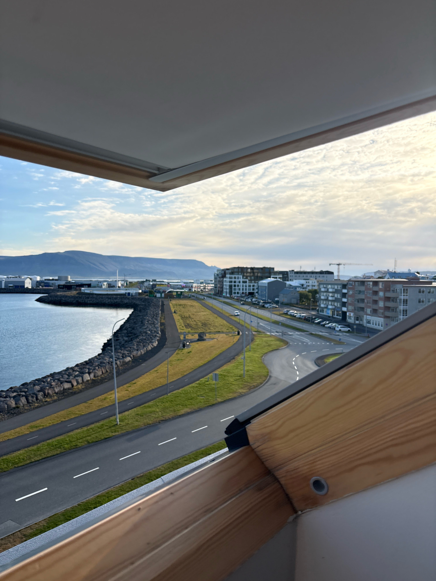 The image depicts a view from an open skylight looking out over a coastal road and cityscape in Reykjavik, Iceland. In the foreground, wooden beams of the skylight frame the view. Beyond the skylight, a dark, rocky breakwater lines the edge of a body of calm, blue water. A paved road with white lane markings curves along the coastline, bordered by green grass. In the mid-ground, a series of modern buildings, including apartment blocks and commercial structures, are visible. A construction crane can be seen among the buildings. Further in the distance, a range of low, blue mountains forms the horizon under a partly cloudy sky. Cars are parked along the road and a few are in motion. The scene appears to be set during daylight hours.