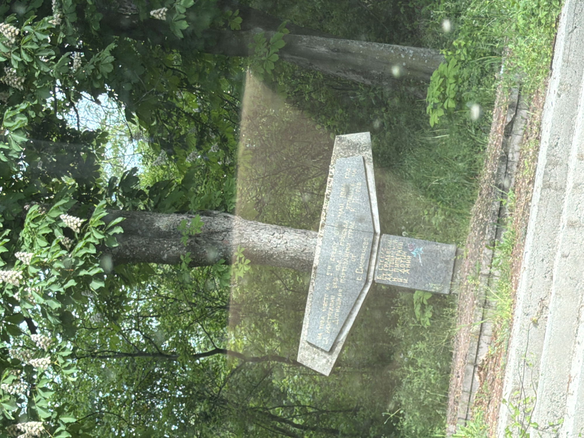 The image shows a stone memorial in a green, outdoor setting. The memorial is cross-shaped and has Cyrillic inscriptions, which are partially obscured. The memorial reads, "HA TOBA MЯCTO HA БИ СТРАДА ЯТАЦИТЕ СA PA3CTРЕЛЯНИ БЕЗ БЕЗ СТАНА ПРИСЪДАН ОТ ДУПНИШКИЯ ПАРТИЗАНСКИ ОТ С. САМОРАНОВО Б. СТОКИС, СТ. ГРАДЕВСКИ, АЛ.СТОИМЕНОВ, H. ДОТИИ, и. Анин." The area surrounding the memorial is filled with trees and vegetation. There are stone steps to the side. The location is likely in or near Blagoevgrad, Bulgaria, based on the Cyrillic text. The memorial appears to commemorate locals with partisan affiliations killed in or near the village of Samoranovo.