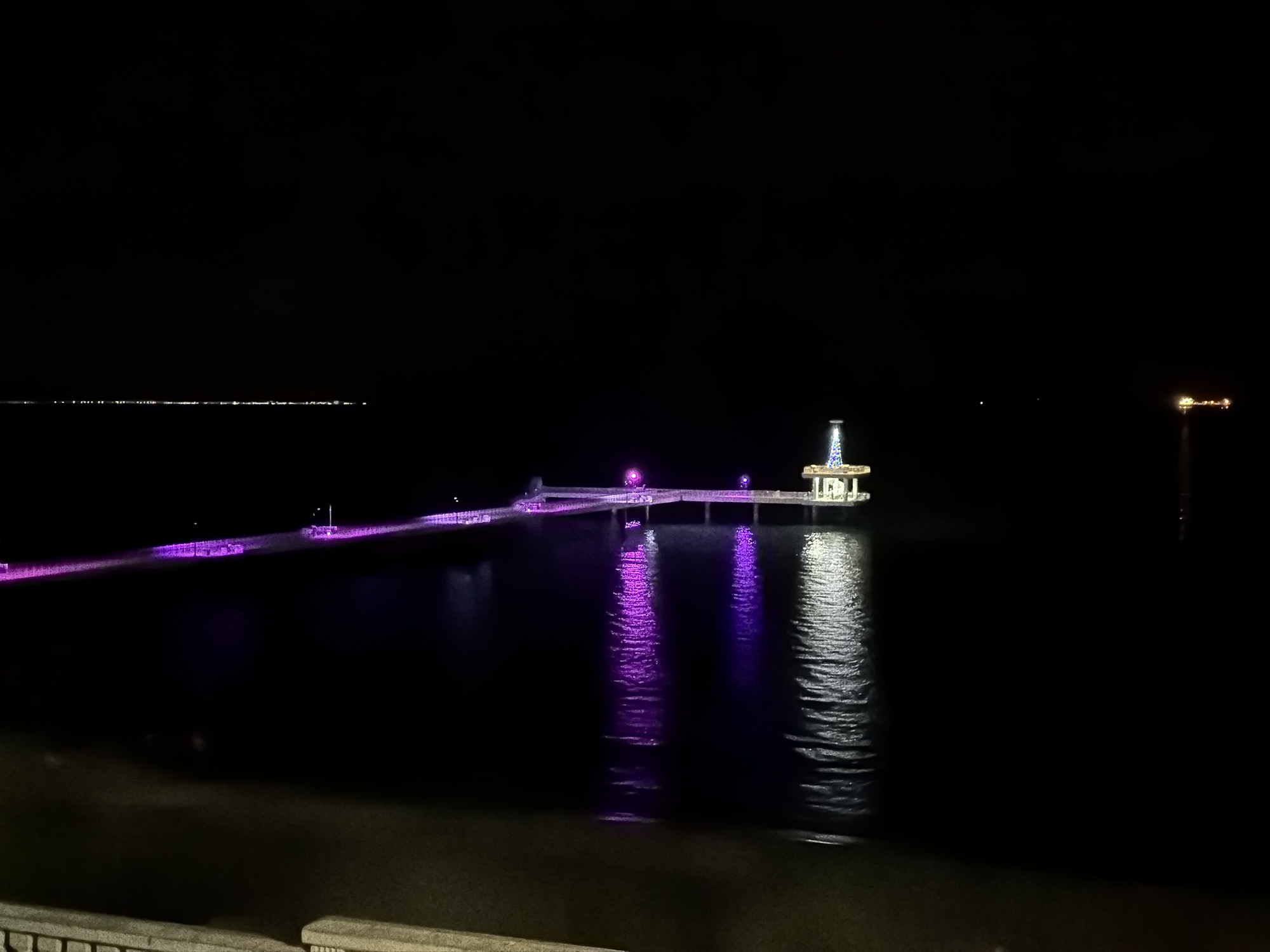At night, a long, brightly lit pier extends into the dark water. The pier is lined with evenly spaced purple lights. At the end of the pier is a structure with a central spire illuminated with white light. The pier and building are reflected in the water. A few faint lights are visible on the horizon. A tall object with dim lights is on the right side of the image. The scene is in Burgas, Bulgaria, specifically on the pier.