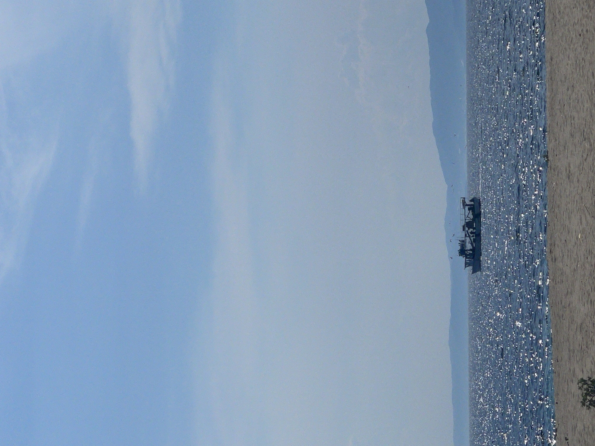 An overview of the Aegean Sea and a boat. The image is a medium shot. The water is a deep blue, sparkling with sunlight. A boat with a white cabin is in the distance. Mountains are visible on the horizon. The sky is a light blue with sparse white clouds. The foreground is a sandy beach with a small bush. The scene is tranquil and serene.