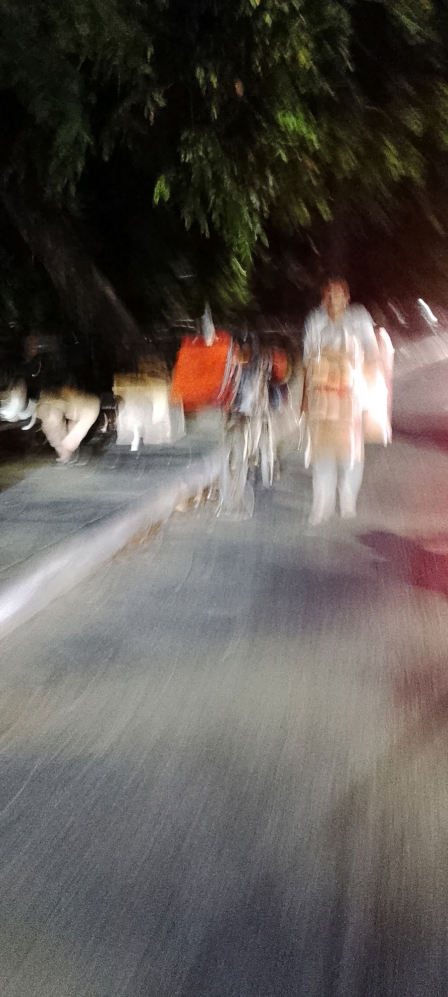 An abstract, motion-blurred nighttime photograph taken on a paved road in Bekasi, Indonesia, depicts several figures and objects. In the background, dark foliage suggests trees lining the street. In the mid-ground, a person wearing light-colored clothing, possibly a traditional garment, stands to the right. To the left of this figure, there are blurred shapes of people and a bright orange object, likely a piece of fabric or sheeting. A bicycle with blurred wheels is positioned centrally. The road surface is visible in the foreground, with a light streak suggesting movement or reflection. The overall impression is one of hurried activity or a long exposure capture during a low-light condition.