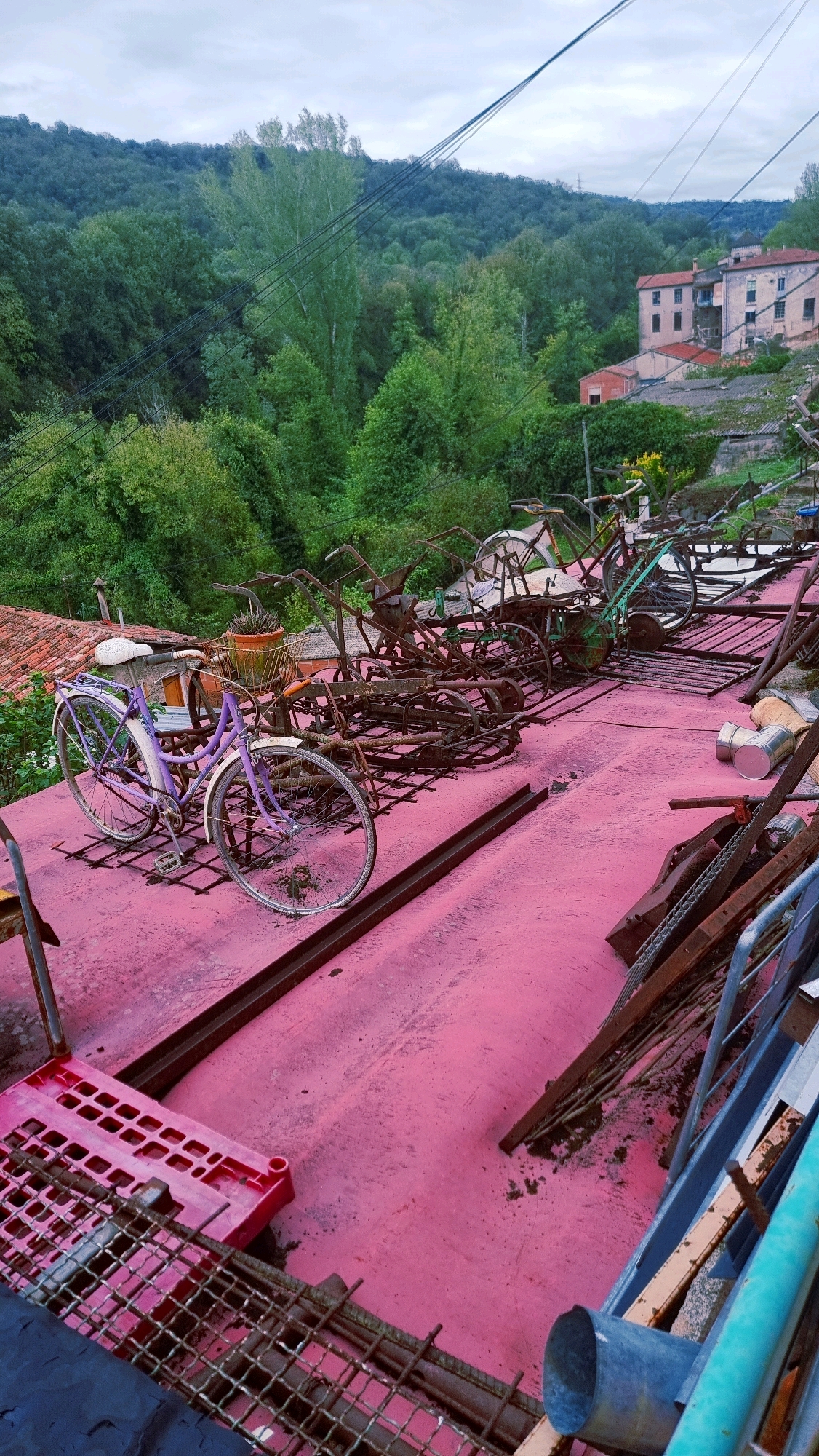 In La Canya, Spain, a cluttered, outdoor scene is depicted on what appears to be a rooftop or elevated platform. The surface is painted a vibrant pink and is covered with various items. In the foreground, a light purple bicycle with a white seat is parked. Behind it, a collection of rusty, old agricultural implements and bicycles are piled together, suggesting a storage or disposal area. The background shows a dense, green forest covering a hillside, with a few buildings visible amongst the trees. The sky is overcast, indicating a cloudy day. The image captures a mixture of old machinery, discarded items, and a natural landscape.
