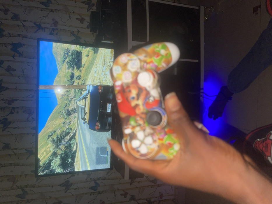 A person holds a custom-designed game controller, featuring cartoon character graphics, in front of a large screen displaying a video game scene. The game shows a black car driving on a road amidst mountainous terrain under a clear blue sky. The background includes furniture and walls with patterned wallpaper. A separate figure partially visible has dark clothing and gloves. Blue lighting emanates from behind the furniture.
