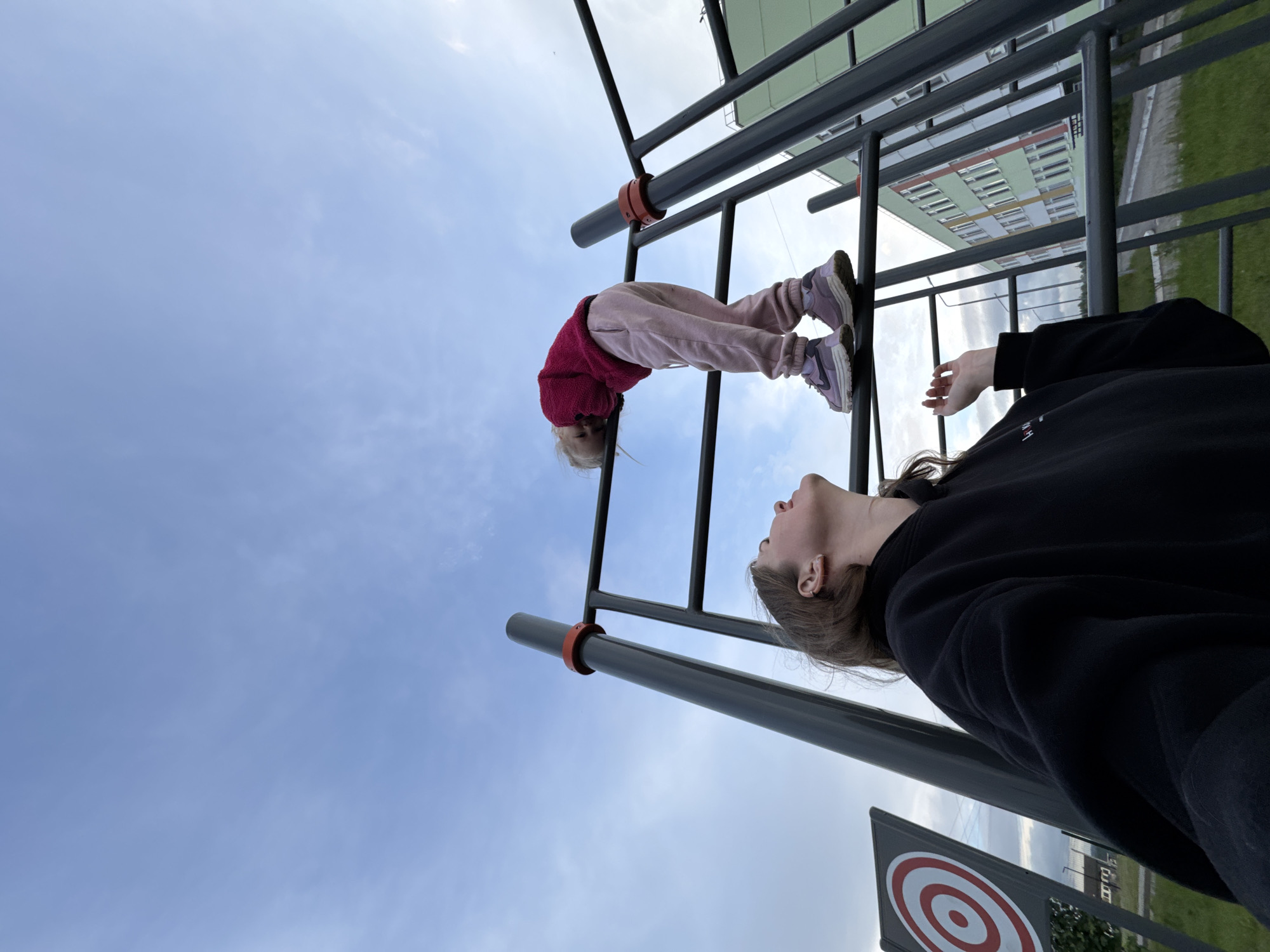 The image displays an outdoor scene featuring two individuals and a metal playground or exercise structure under a partly cloudy, light blue sky. In the lower-left foreground, an adult or teenager, with light brown hair and wearing a black hoodie, is looking upward, their right hand slightly raised with fingers loosely curled. Higher on the grey metal structure, a child wearing a dark pink long-sleeved top, light pink sweatpants, and light purple athletic shoes (Nike branding visible on the left shoe) is leaning forward over a horizontal bar. The metal structure incorporates orange-red rings on its vertical posts. To the left, a grey target board with red and white concentric circles is partially visible. In the mid-ground and background, a multi-story building with a light green facade, numerous windows, and white and orange horizontal stripe details is prominent. Green grass covers the ground in sections, and a paved area is visible. The scene is consistent with an urban recreational area, specifically located in the city of Горный, Russia.