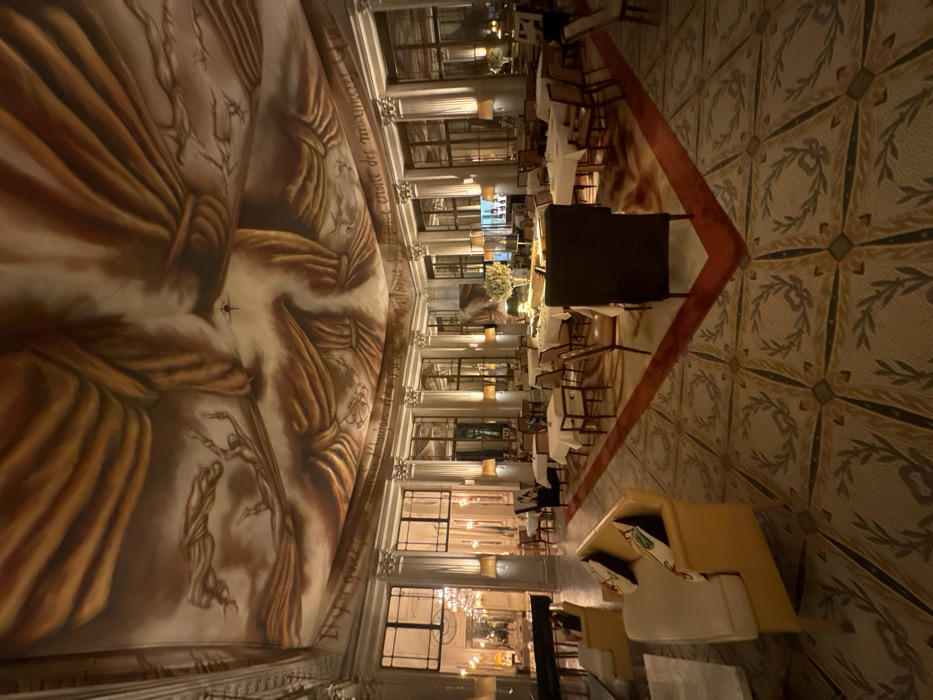 The image displays the lavish interior of Le Dali restaurant, located within the Hôtel Le Meurice on Rue de Rivoli in the 1st arrondissement of Paris, France. The room is spacious and dimly lit, featuring a prominent, expansive ceiling mural rendered in warm ochre, brown, and cream tones, depicting large draped fabric forms, ethereal cloud-like elements, and several abstract human figures in various states of suspension or fall. A partial inscription in cursive script is visible along the mural's lower edge.
The walls are architecturally classical, adorned with numerous regularly spaced fluted columns topped with ornate capitals. Between these columns are large, multi-pane windows that reflect the interior and suggest an adjacent space or exterior. Illumination is provided by multiple wall-mounted sconces with cream-colored shades, casting a warm glow, and ambient light sources reflecting off the ceiling.
The floor is a decorative mosaic or tile pattern, primarily in muted greens, creams, and grays, featuring repeating square panels each containing a laurel wreath motif. A distinct, dark red border delineates a central dining area.
The furniture includes numerous dining tables, mostly rectangular and square, uniformly covered with white tablecloths and surrounded by a variety of chairs with dark frames and light-colored upholstered seats. In the left foreground, two mustard-yellow upholstered couches with cream seat cushions and patterned throw pillows are positioned near a dark, reflective surface, possibly a grand piano. A large central table in the mid-right features a prominent white floral arrangement and other serving items. The room appears to be empty of individuals.